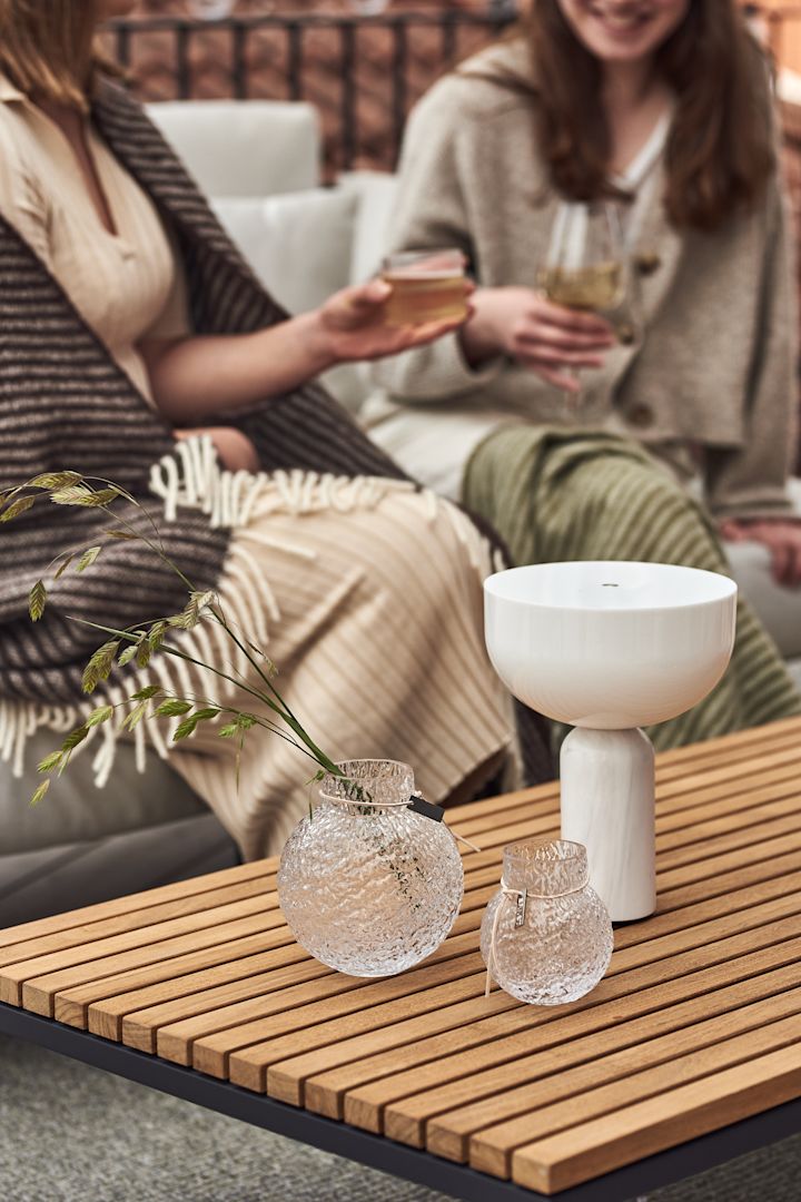 Zwei Frauen sitzen auf einem Sofa auf dem Balkon und stoßen an. Im Vordergrund stehen die kabellose Kizu Tischleuchte von New Works und Vasen von Ernst in verschiedenen Größen auf einem Holztisch.