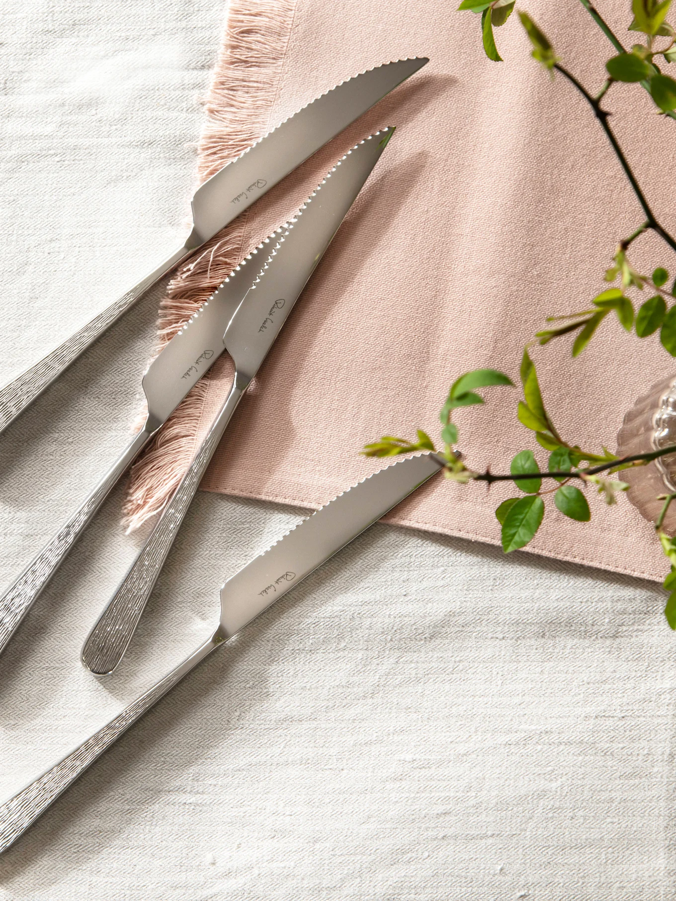 Vier Steakmesser aus Edelstahl mit gezackten Klingen und strukturierten Griffen auf einem Leinentischtuch mit einer rosa Serviette.