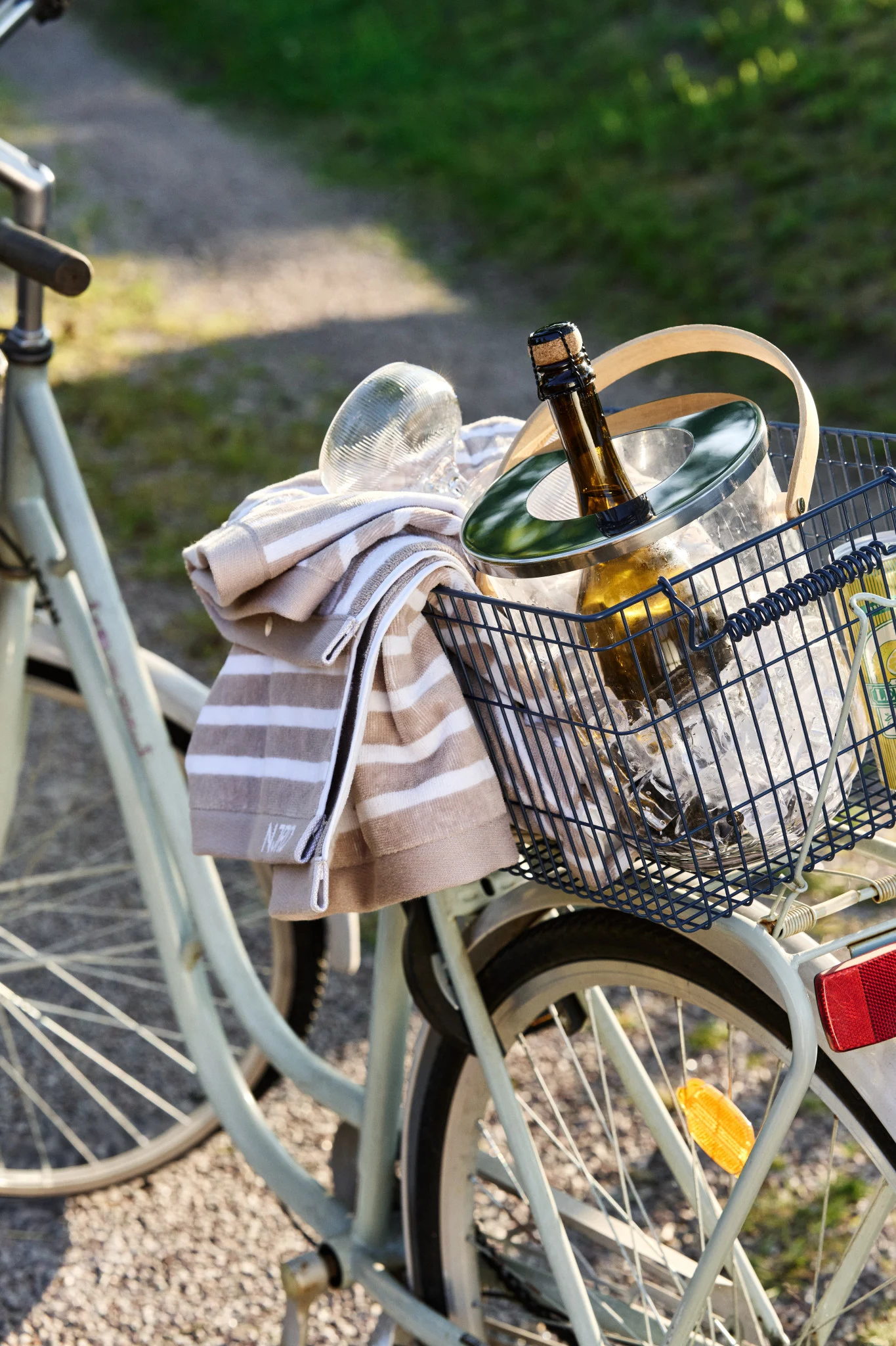 Ein Fahrradkorb enthält einen Eiskübel mit einer kühlenden Flasche und einem gestreiften Handtuch an einem sonnigen Tag.