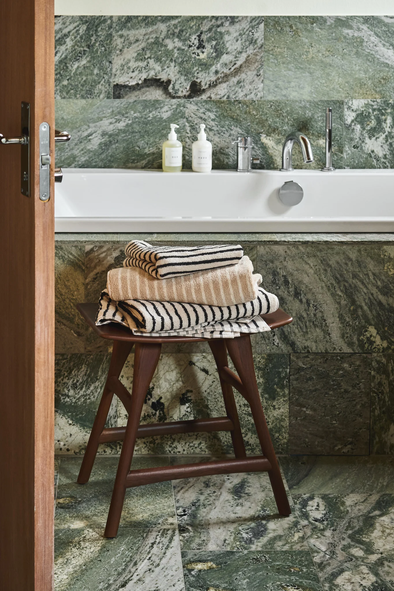 Badezimmer mit grünen Marmorfliesen, weißer Badewanne und einem Holzschemel mit beige und cremefarbenen gestreiften Handtüchern.