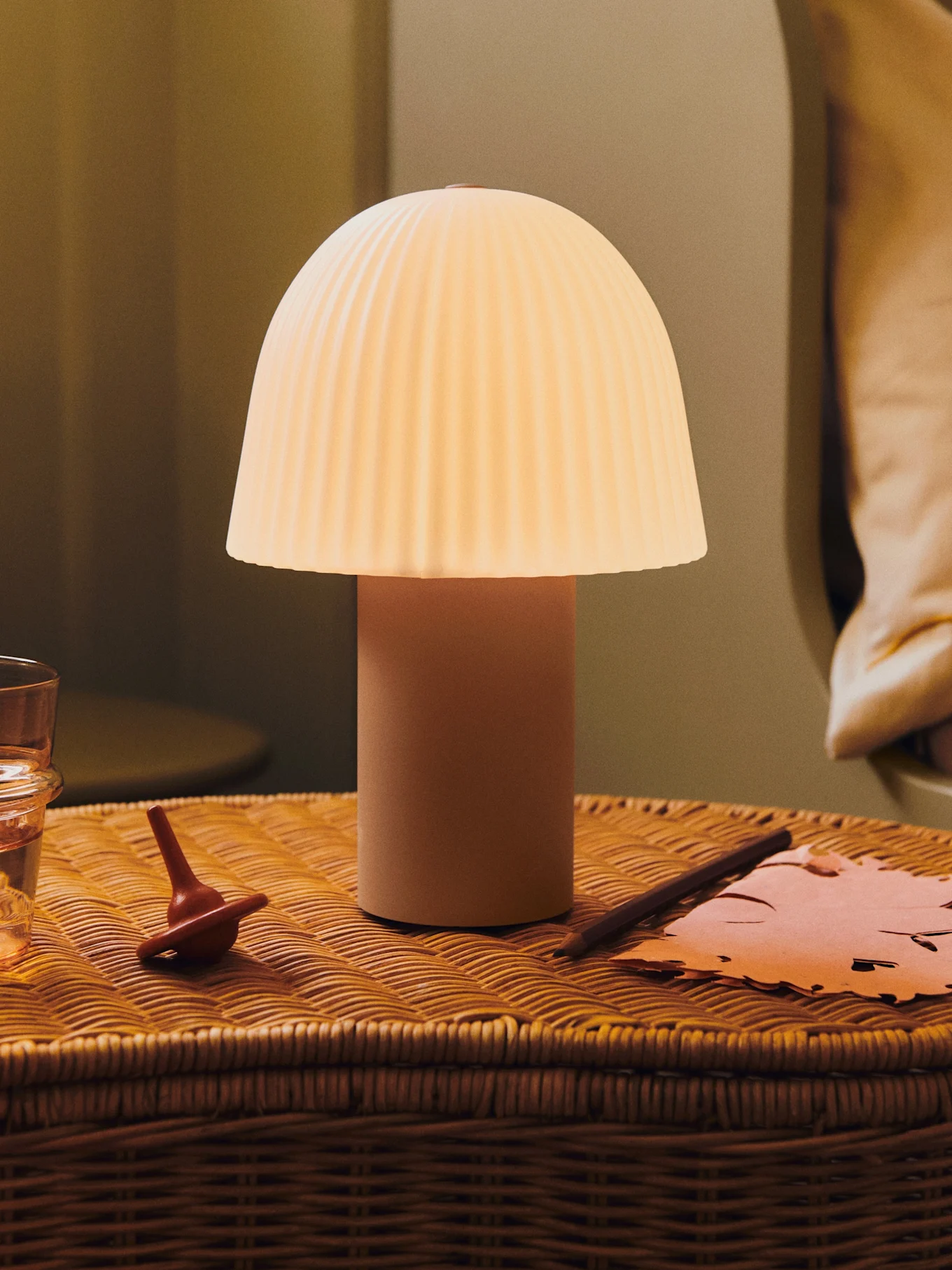 Eine leuchtende Pilzlampe mit plissiertem Schirm auf einem Weiden-Nachttisch mit einem Glas Wasser, einem Kreisel und Papier.