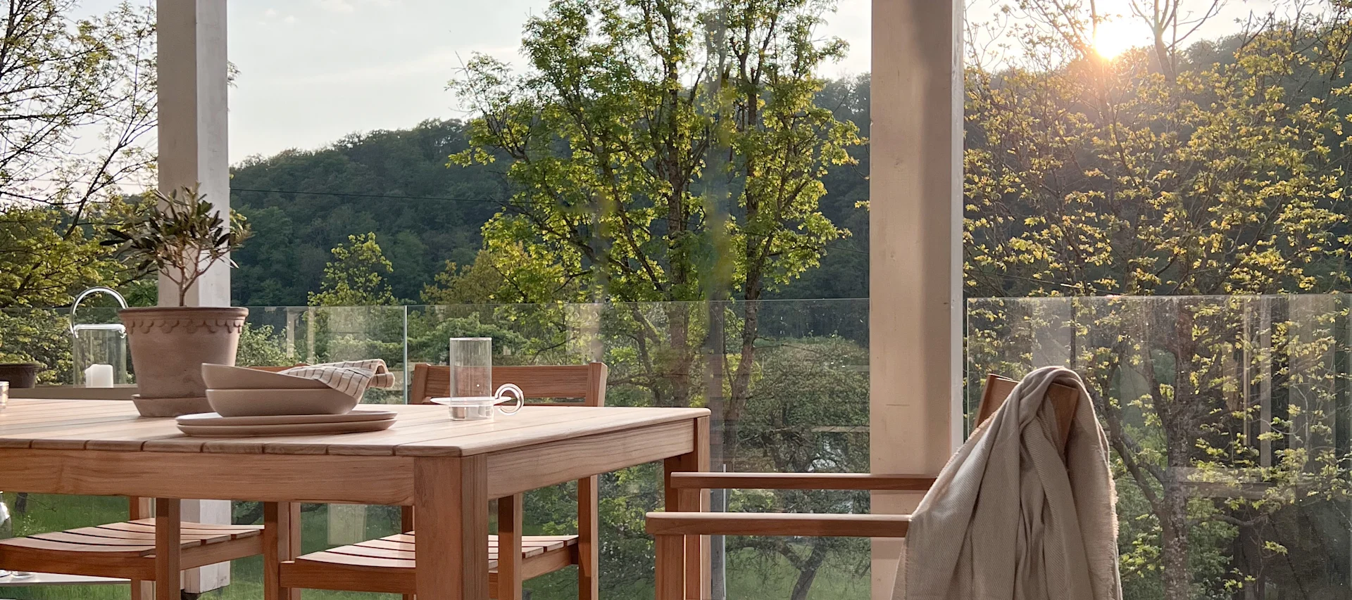 Outdoor-Esstisch aus Holz mit Tellern und Gläsern auf einer Terrasse mit Blick auf grüne Bäume im Sonnenlicht.