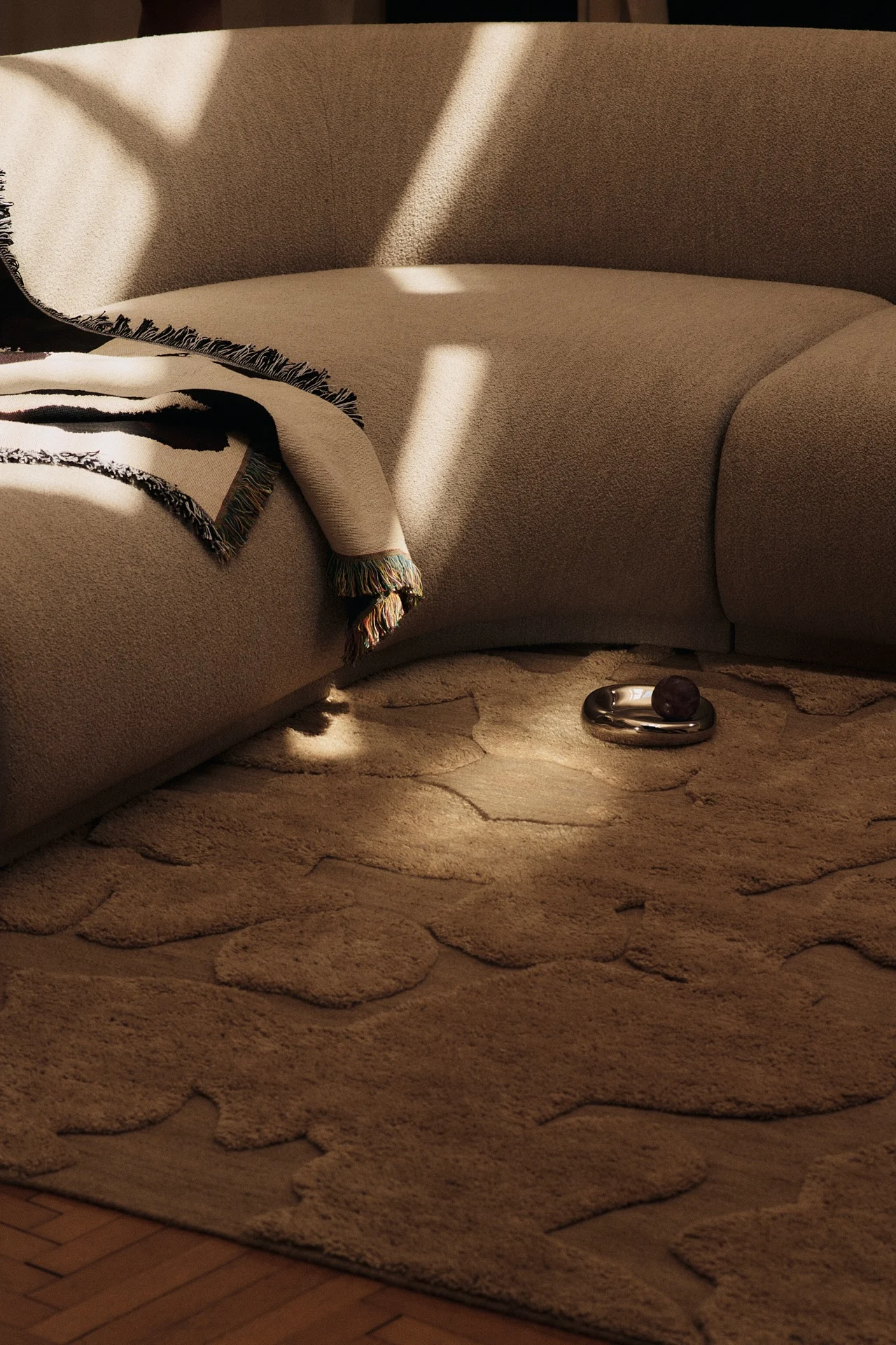 Geschwungenes beiges Sofa mit gemusterter Decke, organisch geformtem beigem Teppich und einer Silberschale mit dunkler Frucht in einem sonnigen Raum.