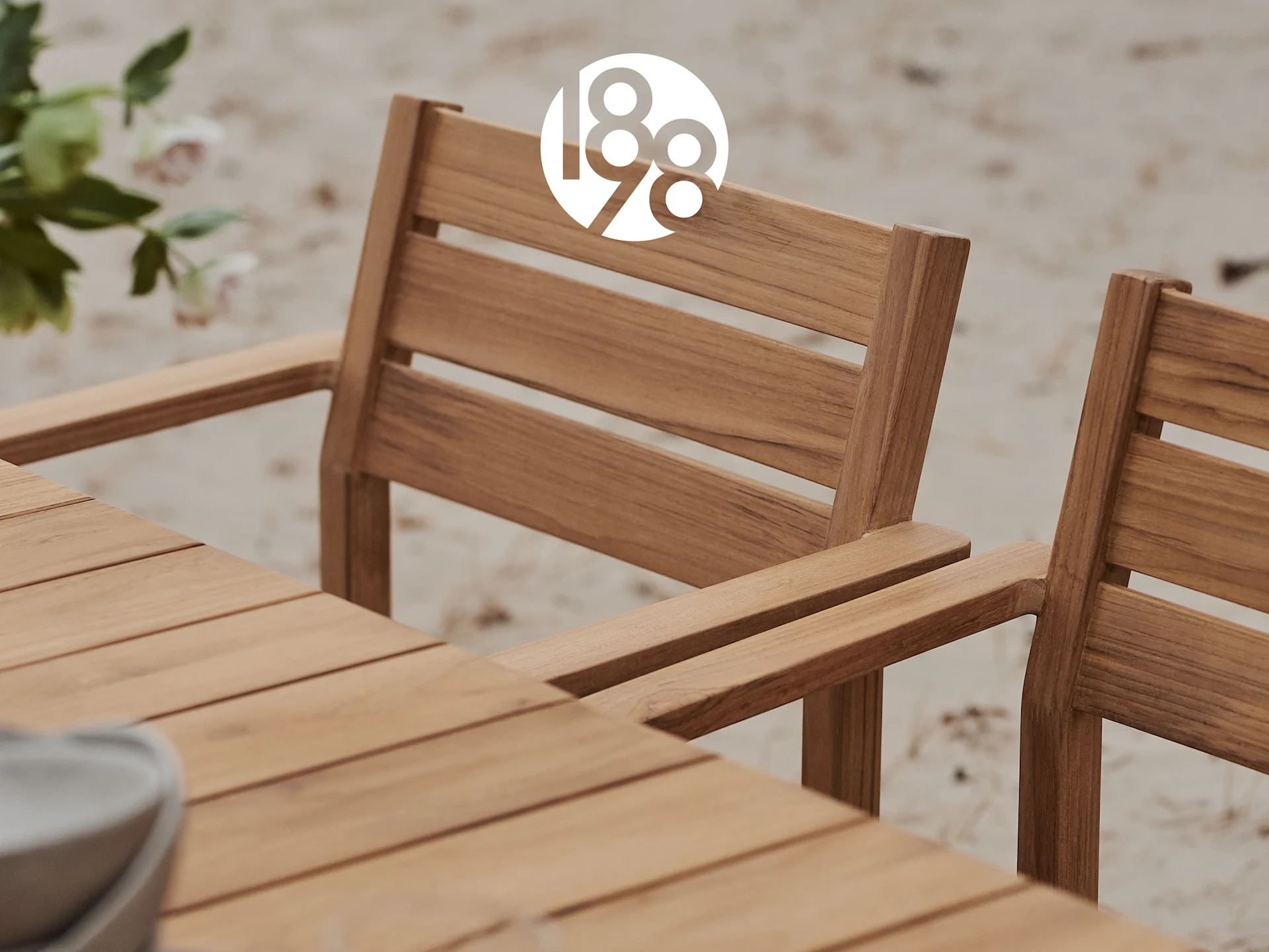 Nahaufnahme eines Esstisches und zweier Stühle aus Holzlatten für den Außenbereich auf Sand, mit einem weißen '188'-Logo.