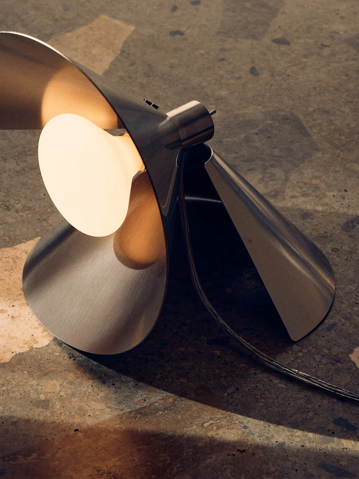 Moderne Tischlampe aus gebürstetem Metall mit skulpturalem, gefaltetem Design und warmer, freiliegender Glühbirne auf Texturfläche.