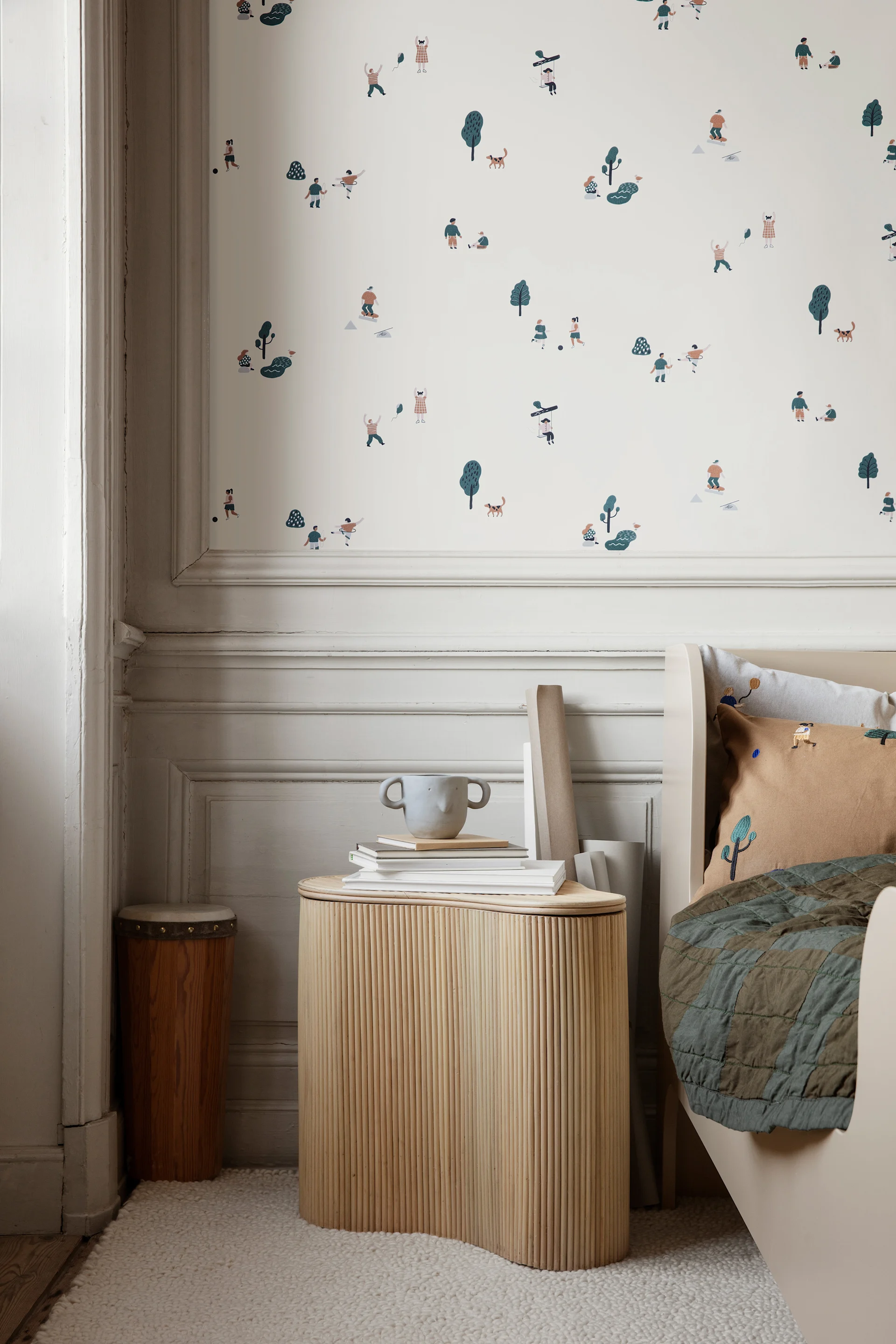 Kinderzimmer mit verspielter Tapete, die Menschen und Tiere darstellt, einem geriffelten Beistelltisch mit Büchern und Tasse sowie einem Bett mit braunem Kissen und grüner Steppdecke.