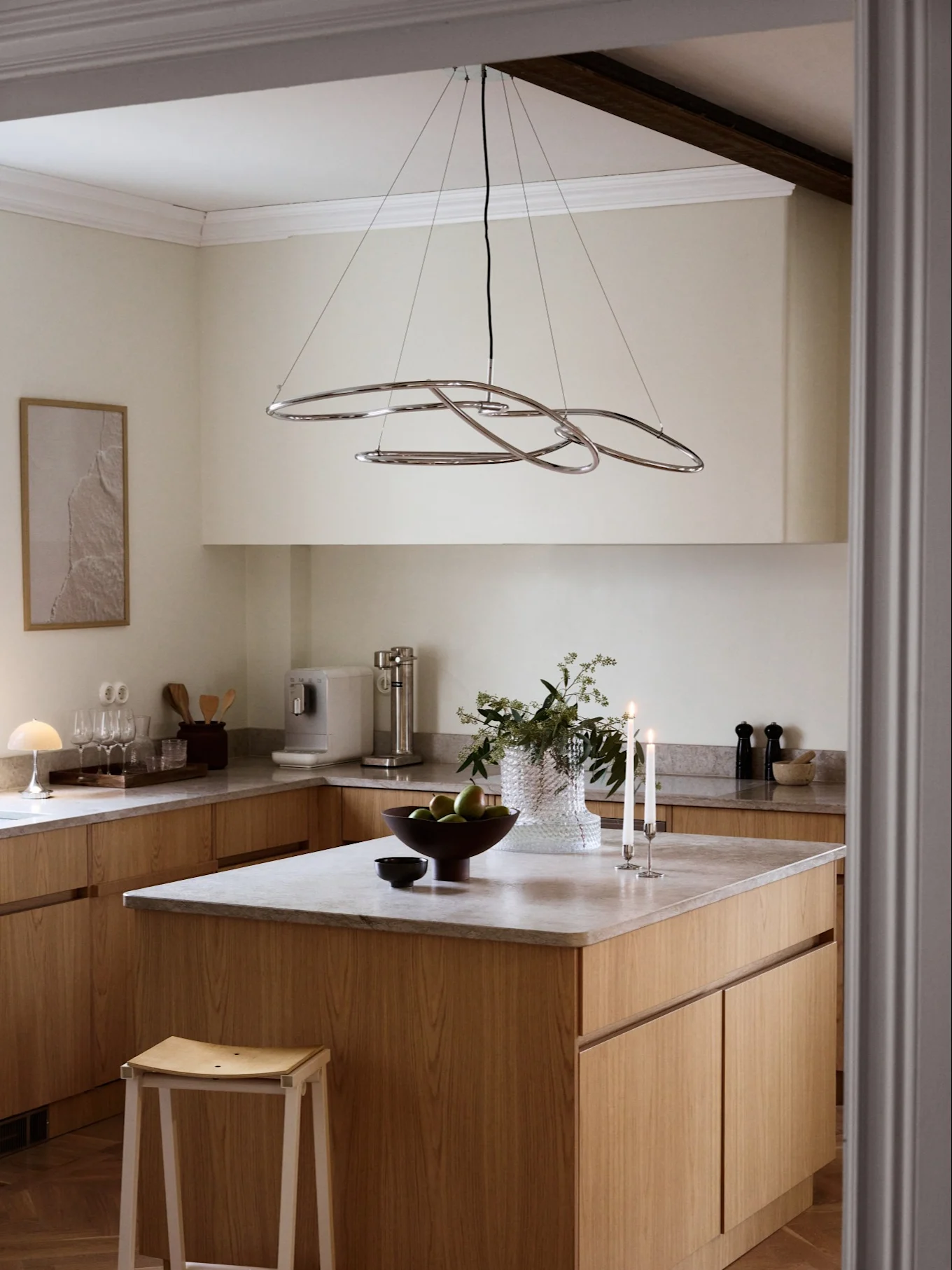 Eine moderne Küche mit hellen Holzschränken, grauen Arbeitsplatten und einem chromfarbenen abstrakten Kronleuchter über einer Insel mit Obst und Kerzen.
