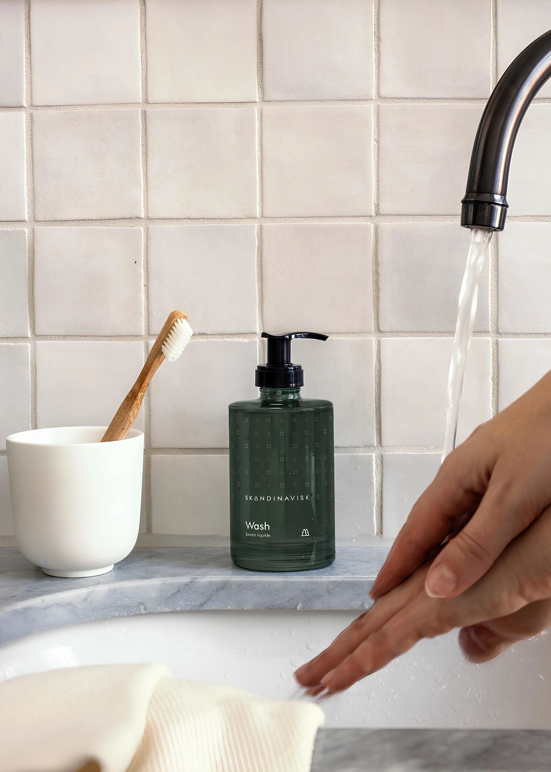 Geschenk zum Valentinstag: Verschenken Sie Spa-Momente. Hier steht die Skog Seife von Skandinavisk auf einem Waschbecken, während sich eine Person die Hände wäscht.