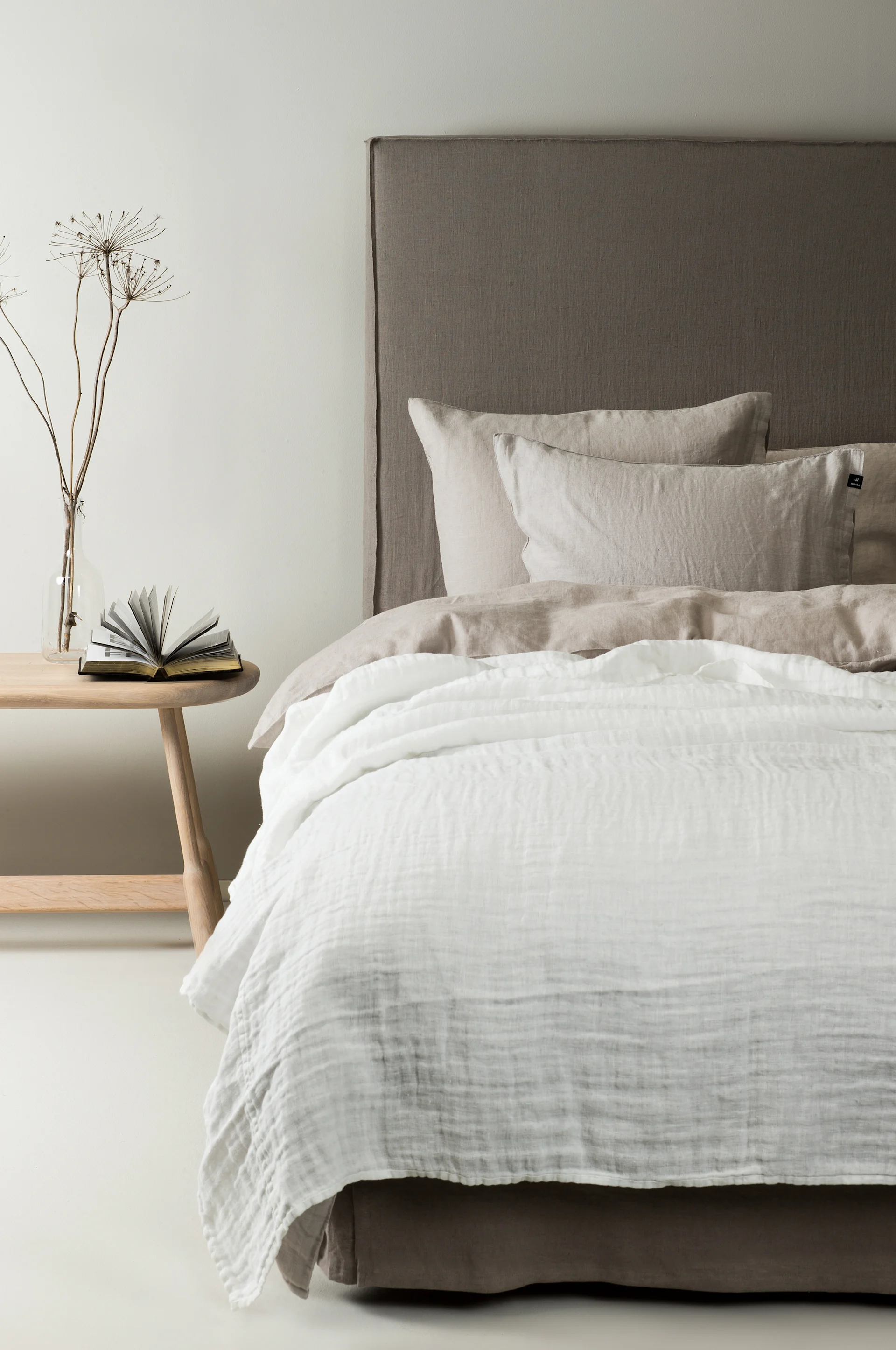 Minimalistisches Schlafzimmer mit Bett, braunem Kopfteil, hellbeiger Leinenbettwäsche, weißer Knitterdecke und Holztisch mit Trockenblumen und offenem Buch.