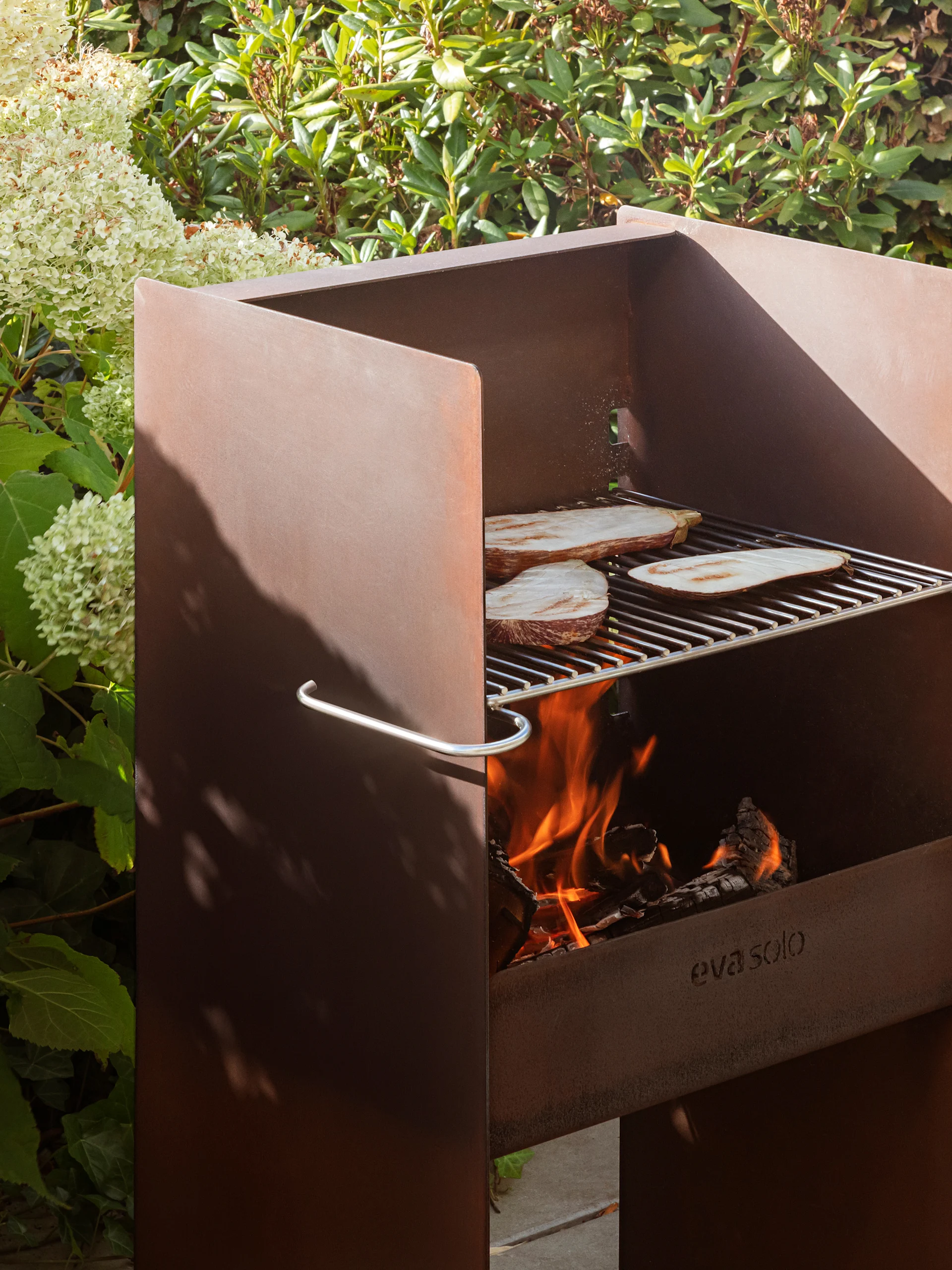 Rostiger Metall-Außengrill von Eva Solo mit Auberginen auf dem Rost, brennendem Feuer und üppigem Laub.