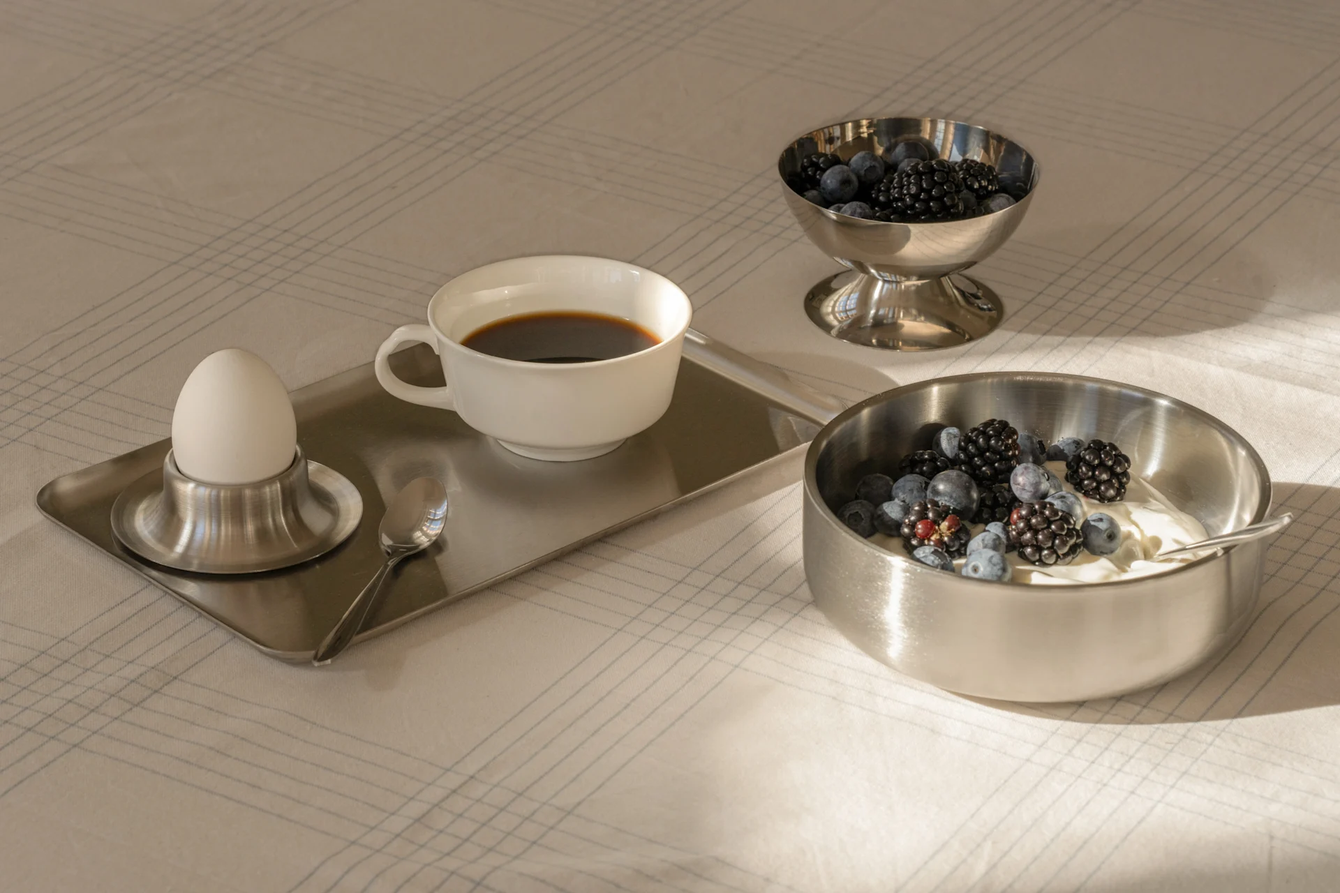 Frühstück mit Kaffee, gekochtem Ei, Joghurt und frischen Beeren in Silberschalen auf einem gemusterten Tischtuch.