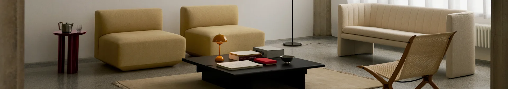 Ein modernes Wohnzimmer mit zwei senffarbenen armlehnenlosen Stühlen, einem schwarzen Couchtisch, einem cremefarbenen gerippten Sofa und einem geflochtenen Holzstuhl.