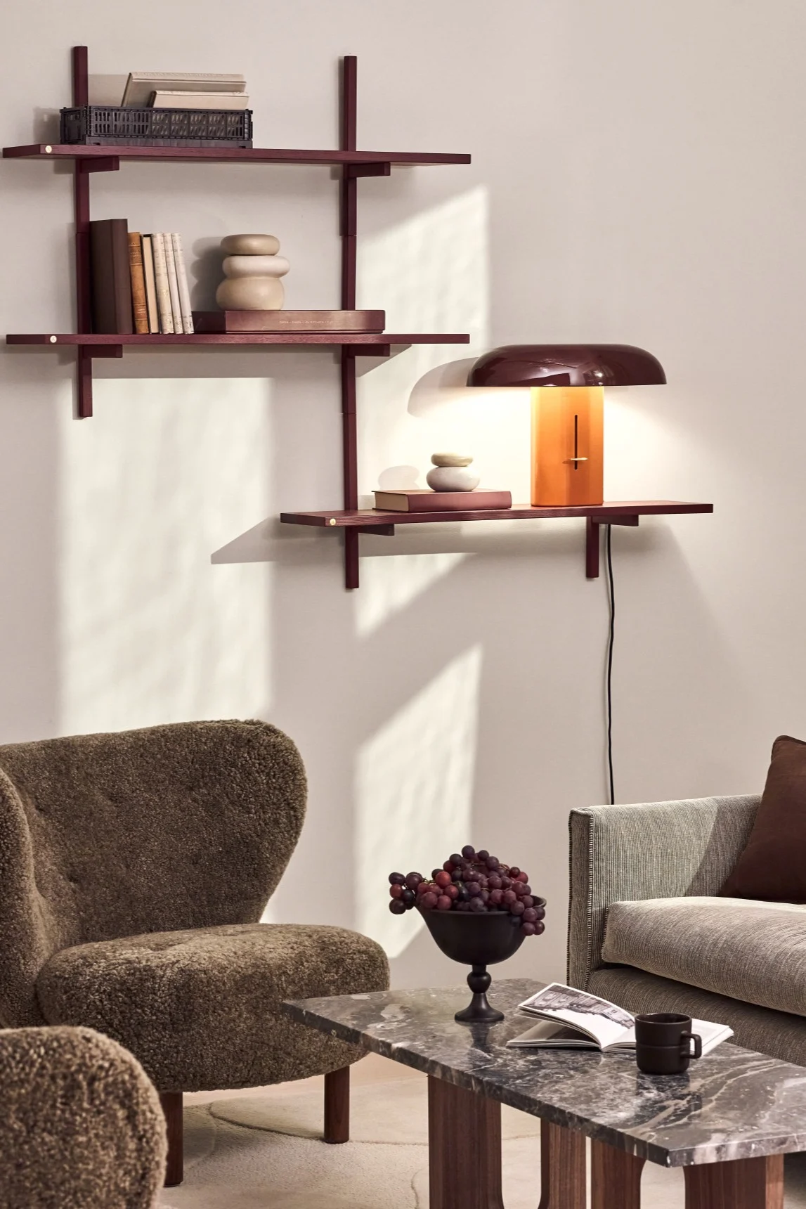 Ein gemütliches Wohnzimmer mit rötlich-braunen Wandregalen, einer Lampe mit gewölbtem Schirm und orangefarbenem Fuß, einem strukturierten braunen Sessel und einem Marmor-Couchtisch mit Trauben und einem Buch.