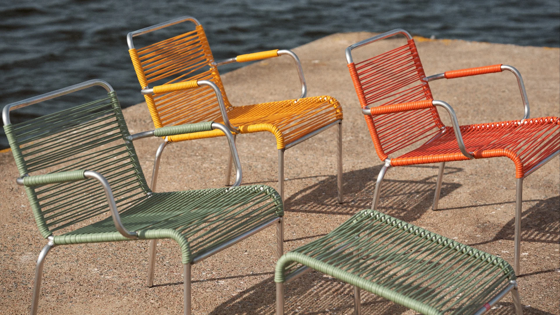 Drei geflochtene Stühle in Grün, Orange und Gelb mit einem passenden grünen Fußhocker auf Sand am Wasser.