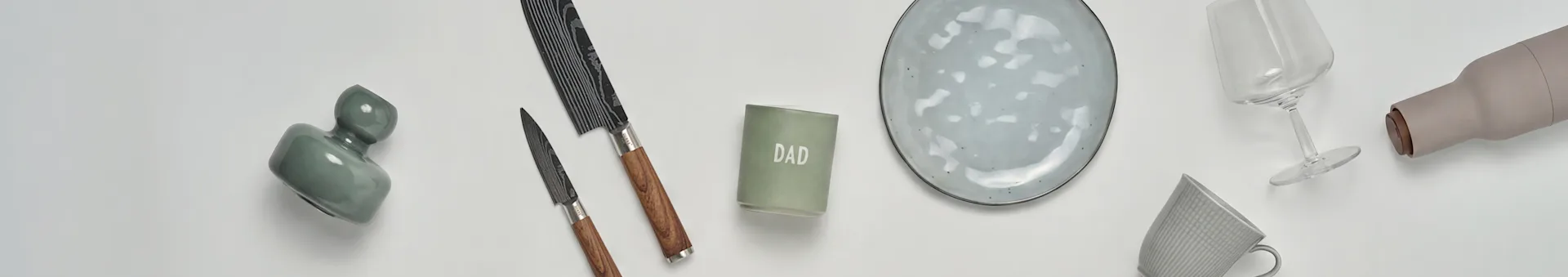 Eine flache Anordnung von Küchen- und Geschenkartikeln: eine grüne Vase, zwei Messer, eine 'DAD'-Kerze, ein grauer Keramikteller, ein Weinglas, eine beige Flasche und eine graue Tasse.