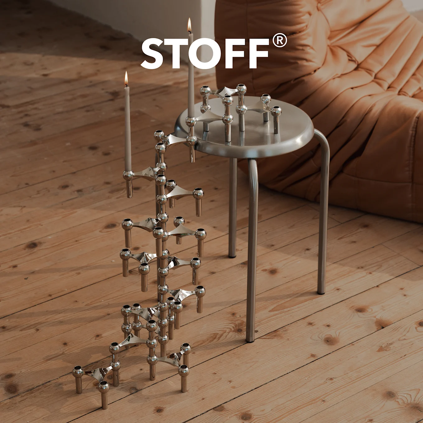 STOFF-Logo über einem Chrom-Kerzenhalter mit brennenden Kerzen auf Holzboden, daneben ein Silberhocker mit einem weiteren Kerzenhalter.
