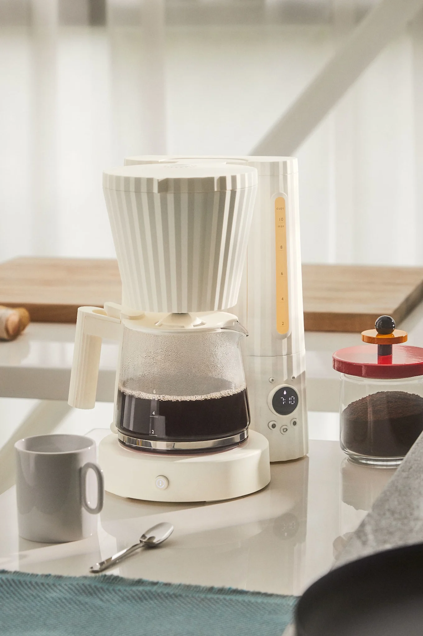 Cremefarbene Filterkaffeemaschine mit Glaskanne voller Kaffee, digitaler Uhrzeit 7:10 und einem Glas mit Kaffeepulver auf einer weißen Arbeitsplatte.