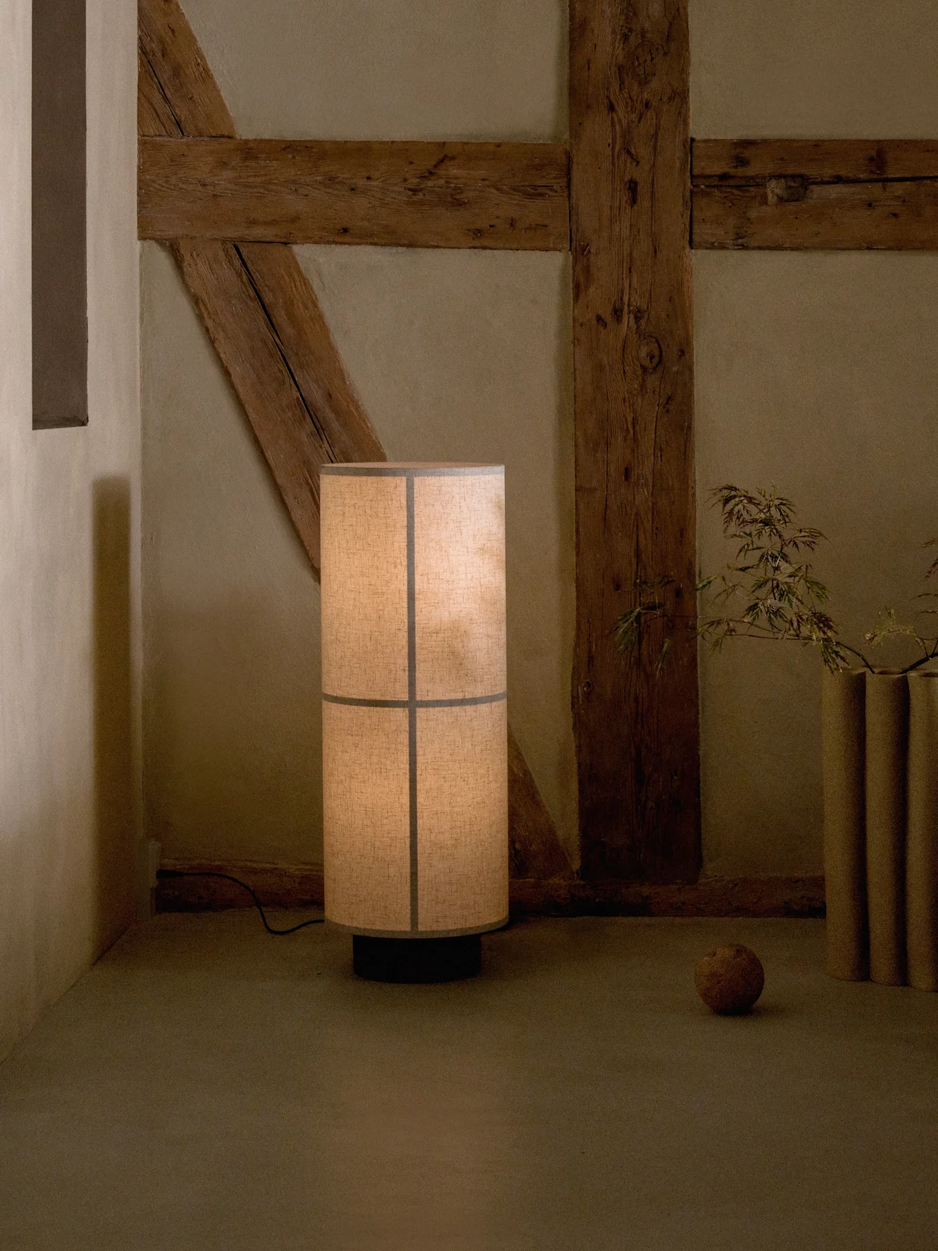 Hohe Stehlampe mit segmentiertem Stoffschirm, die ein warmes Licht in einem rustikalen Raum mit Holzbalken wirft.