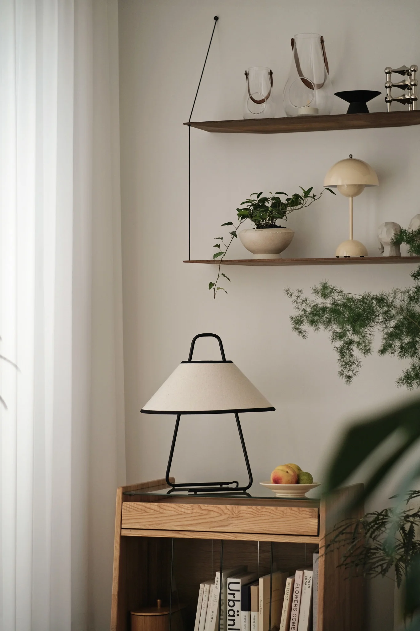 Modernes Wohnzimmer mit cremefarbener und schwarzer Tischlampe auf hellem Holzschrank, Deko-Regalen und Pflanzen.