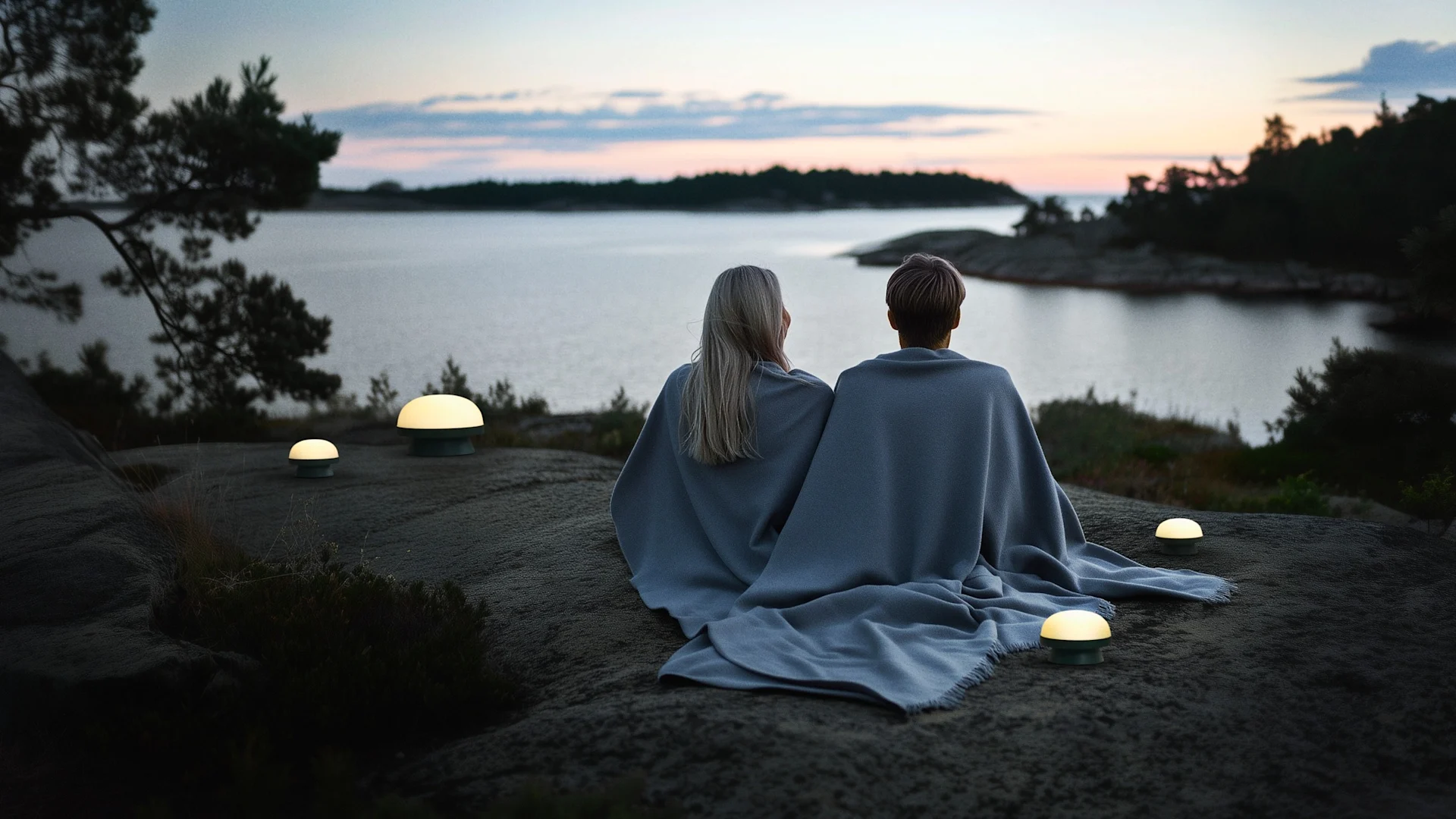 Windlichter & Laternen outdoor: Ein schwedischer See bei Sonnenuntergang mit Felsen davor, auf dem ein Paar mit Decken und mehreren Soft Spot dots Solarleuchten von Rosendahl sitzt.