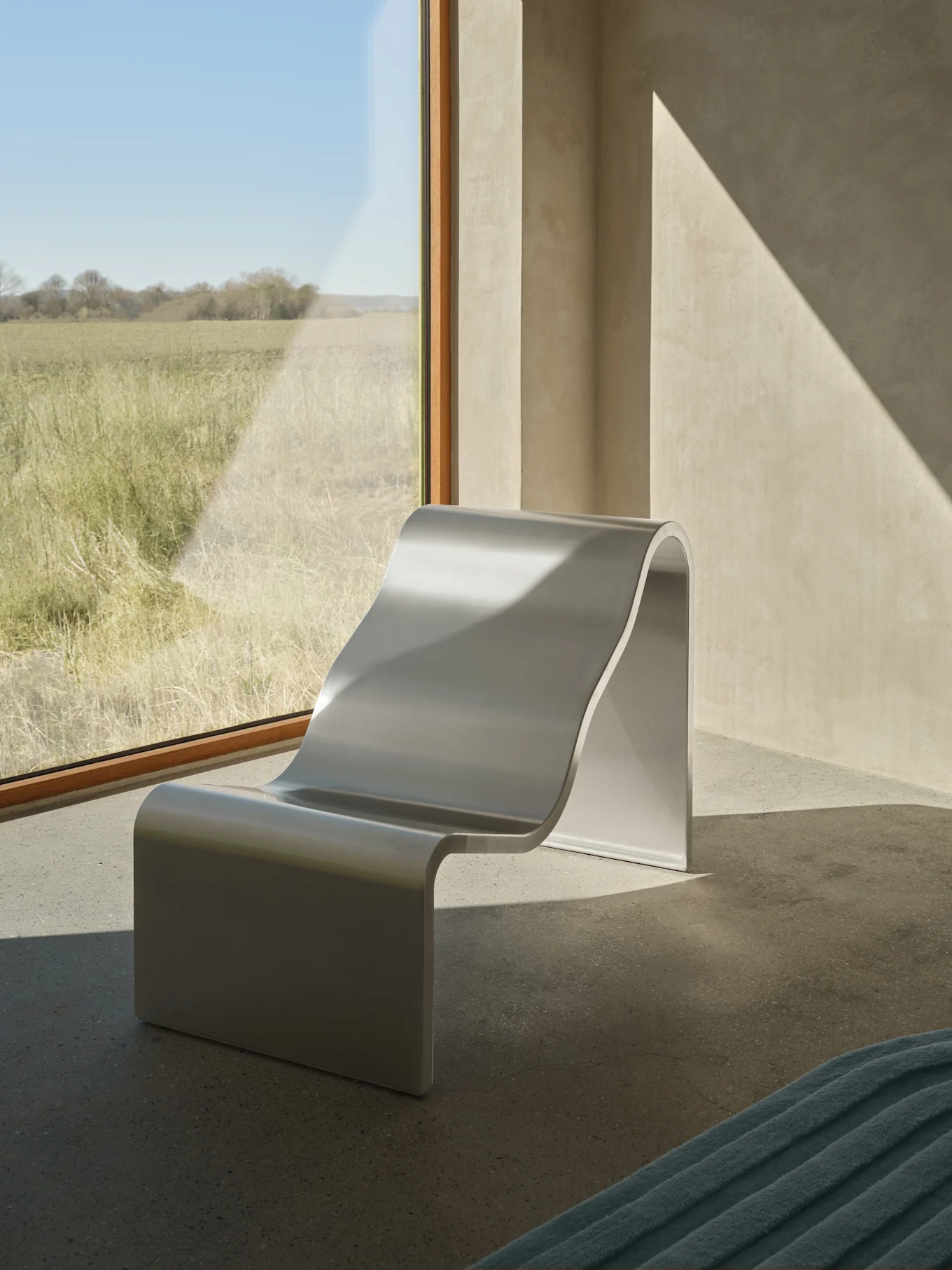 Ein moderner, silberfarbener S-förmiger Loungesessel steht an einem großen Fenster mit Blick auf ein sonniges Grasfeld, mit diagonalen Sonnenlichtmustern an den Wänden.