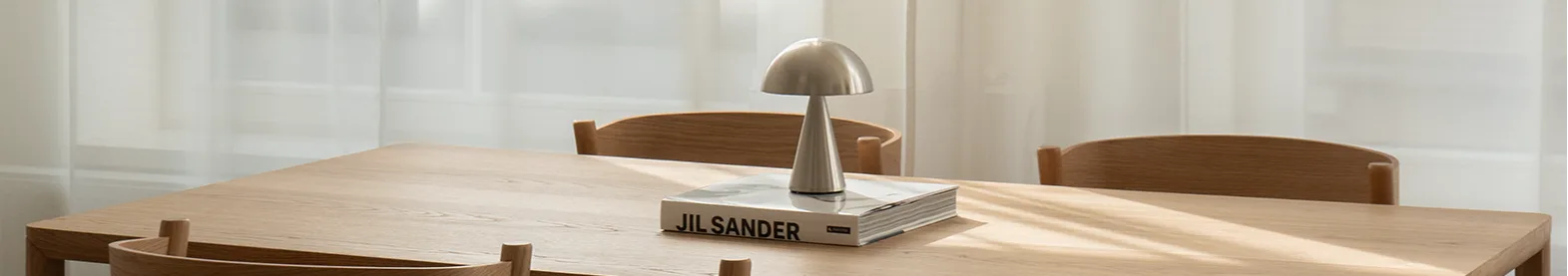 Heller Holzesstisch mit vier Stühlen. Silberne Pilzlampe und Buch auf dem Tisch. Sonnenlicht durch transparente weiße Vorhänge.