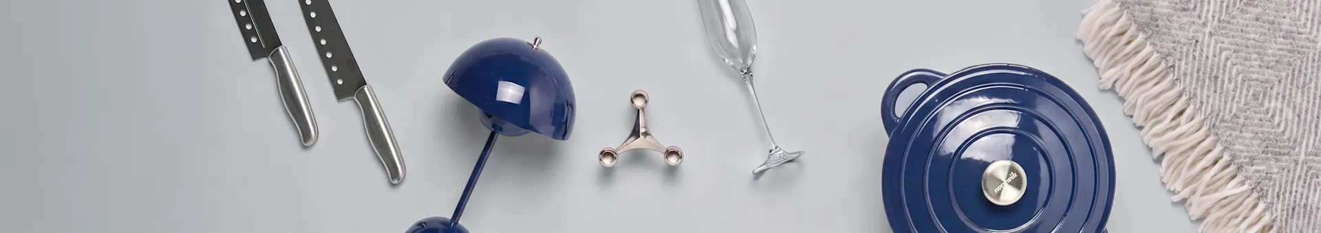Auswahl an Küchenutensilien und Wohnaccessoires: zwei silberne Messer, eine blaue Dekolampe, ein silbernes Küchengerät, ein Sektglas, ein blauer Topf mit Deckel und eine gemusterte Decke.