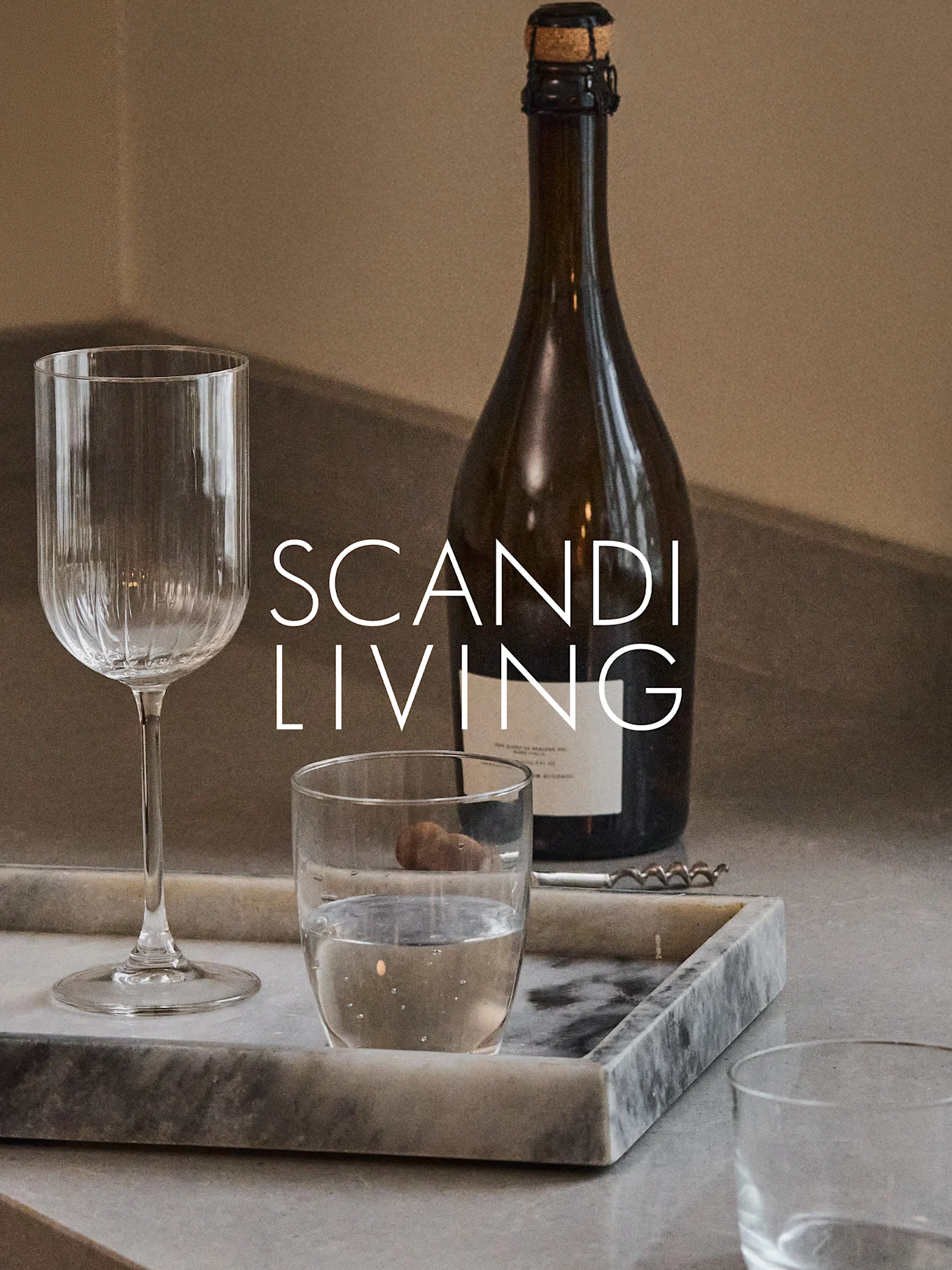 Eine dunkle Flasche, ein geripptes Weinglas, ein Wasserglas und ein Korkenzieher auf einem Marmortablett mit 'SCANDI LIVING' Text.