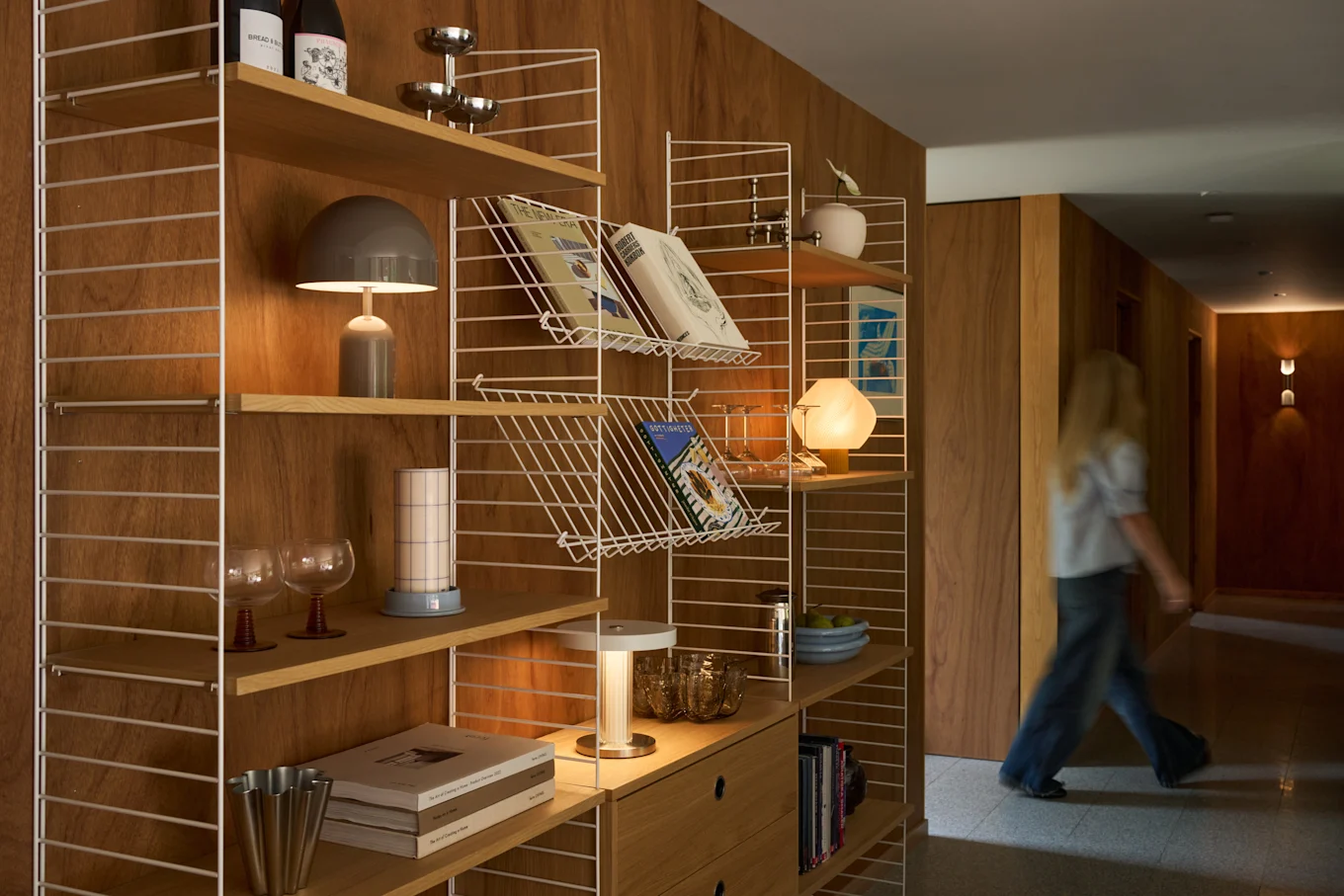 Eine blonde Person geht einen holzvertäfelten Flur entlang, neben einem modernen Regal, das mit Büchern, Lampen und Dekoartikeln gefüllt ist.