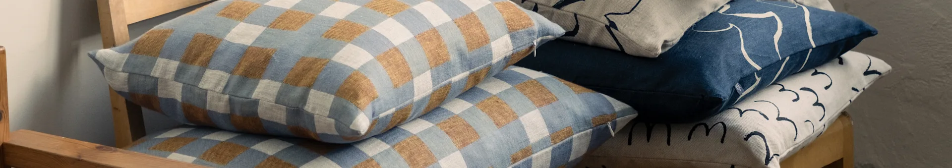 Gestaubte Kissen auf einem Holzstuhl, mit karierten blauen und braunen, sowie abstrakt gemusterten blauen und beigefarbenen Designs.