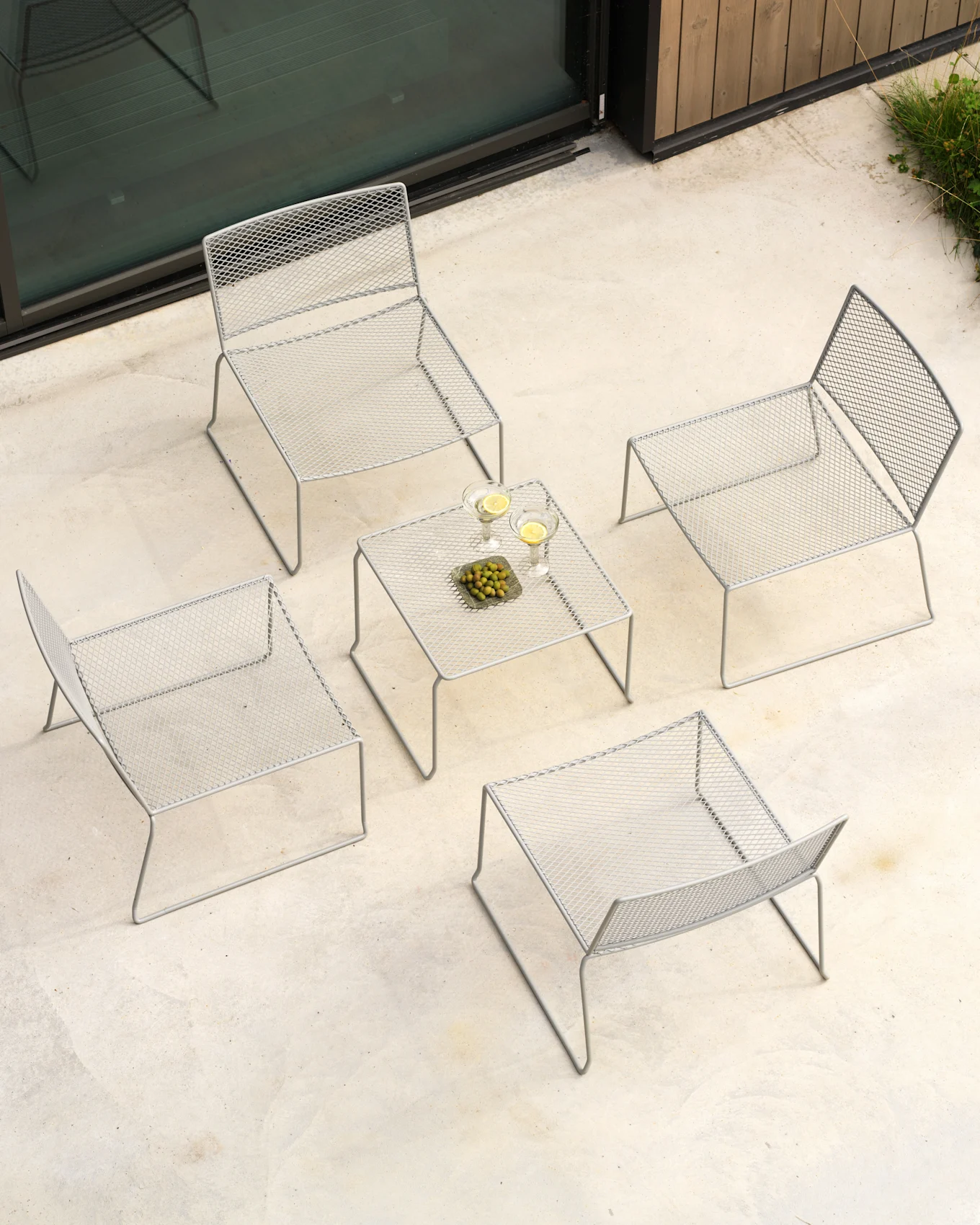 Luftaufnahme eines Outdoor-Terrassensets mit vier grauen Metallgeflechtstühlen und einem passenden Couchtisch mit Getränken und Oliven.