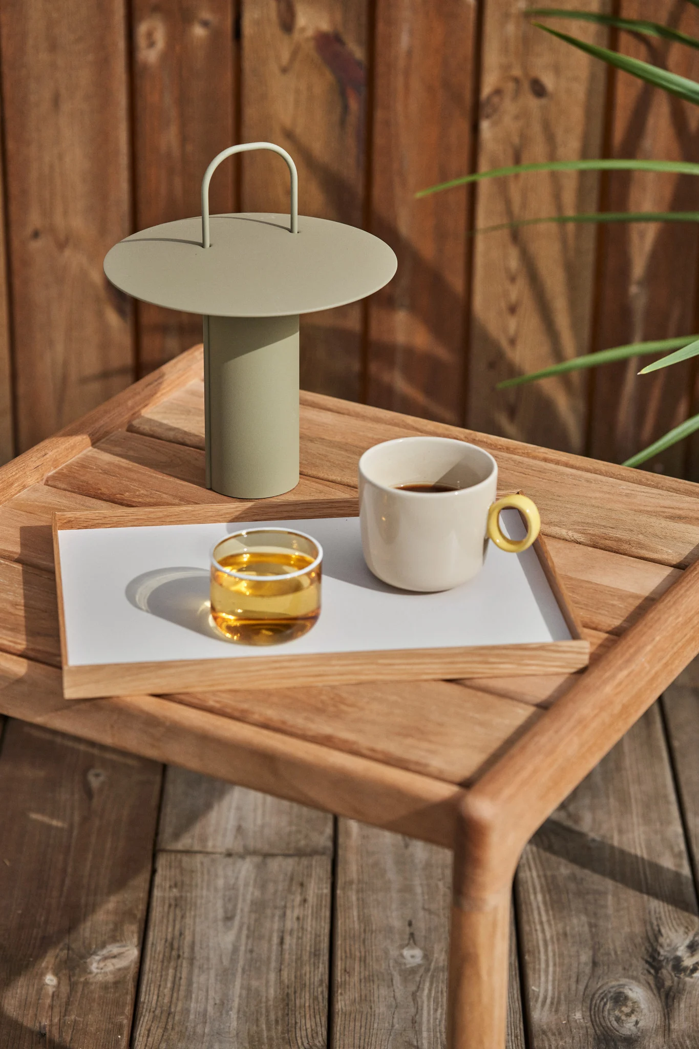 Eine hellgrüne tragbare Lampe, ein Glas bernsteinfarbene Flüssigkeit und eine cremefarbene Kaffeetasse auf einem Holztablett auf einem Holztisch im Freien.