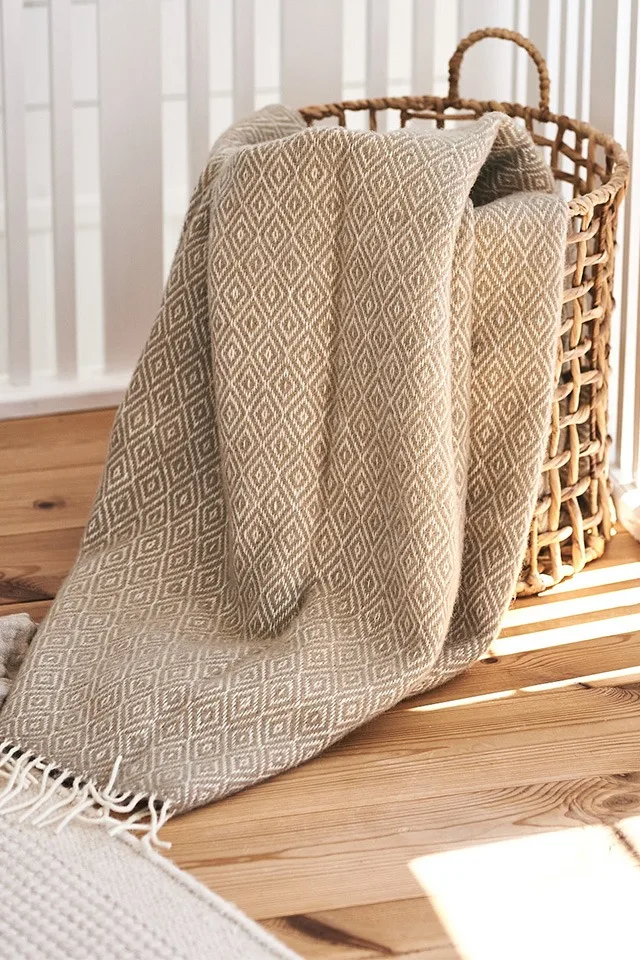 Die beigefarbene Wolldecke Stella von Klippan Yllefabrik liegt in einem geflochtenen Korb von ERNST - bereit für gemütliches Hygge-Vergnügen. 