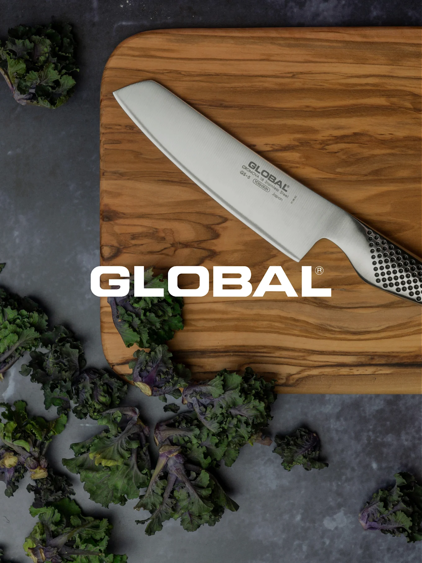 Ein Global-Messer auf einem Holzschneidebrett, umgeben von frischem grünem und lila Blattgemüse.