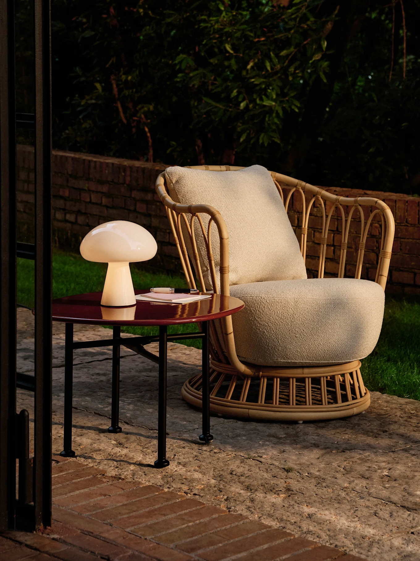 Pilzlampe auf einem roten Beistelltisch neben einem Rattan-Sessel auf einer Terrasse in der Dämmerung.