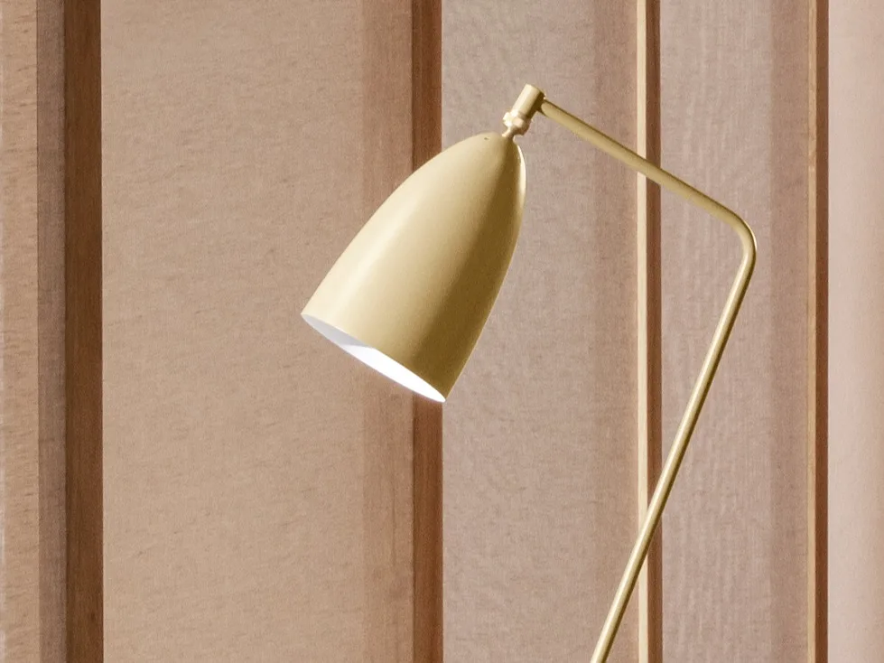 Eine Nahaufnahme einer gelblich-goldenen modernen Lampe mit konischem, nach unten gerichteten Kopf, die Licht vor einer hellbraunen Wandverkleidung abstrahlt.
