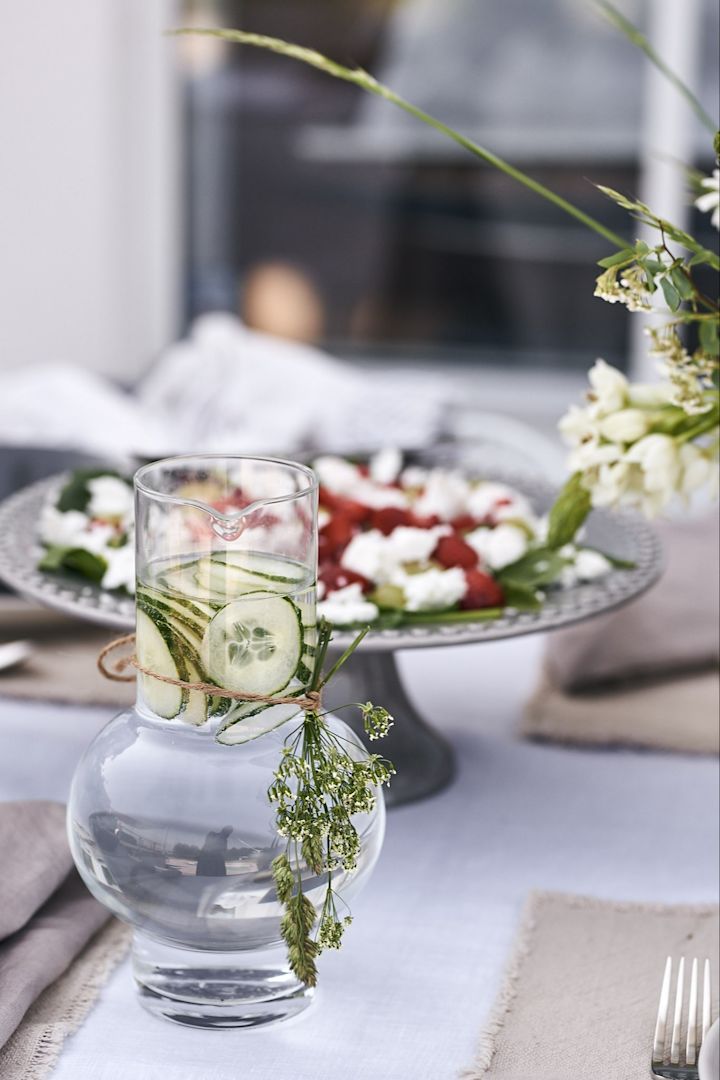 Die stilvolle, mit Wiesenblumen verzierte und mit Wasser und Gurken gefüllte Wasserkaraffe von Ro Collection steht auf dem Tisch. 