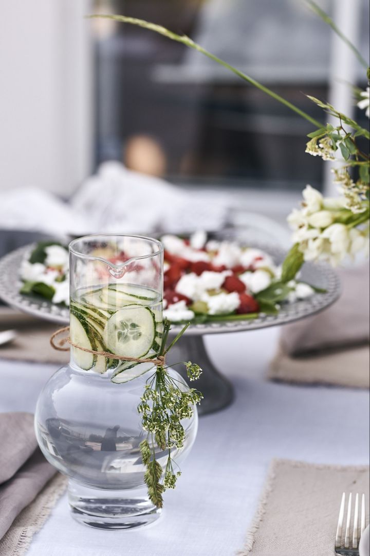 Die stilvolle, mit Wiesenblumen verzierte und mit Wasser und Gurken gefüllte Wasserkaraffe von Ro Collection steht auf dem Tisch. 