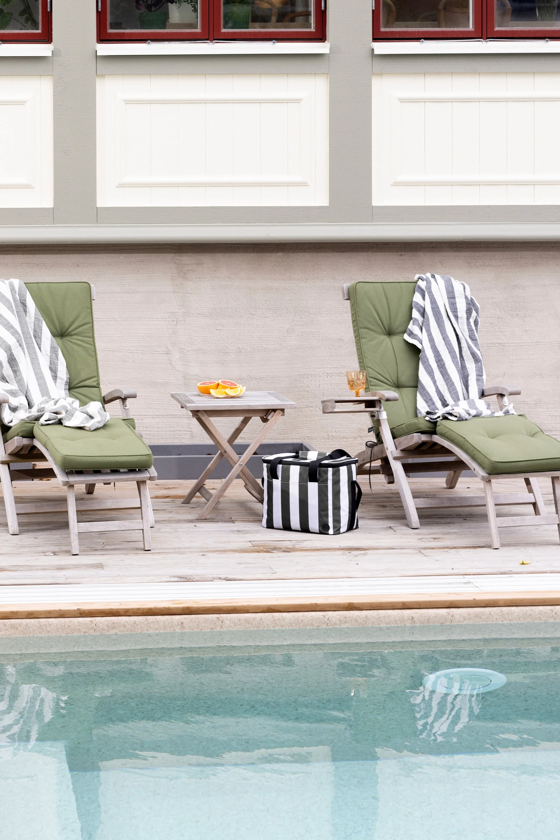 Zwei Holzliegen mit grünen Polstern und gestreiften Decken auf einem Holzdeck am Pool. Eine Kühltasche und Grapefruit sind in der Nähe.