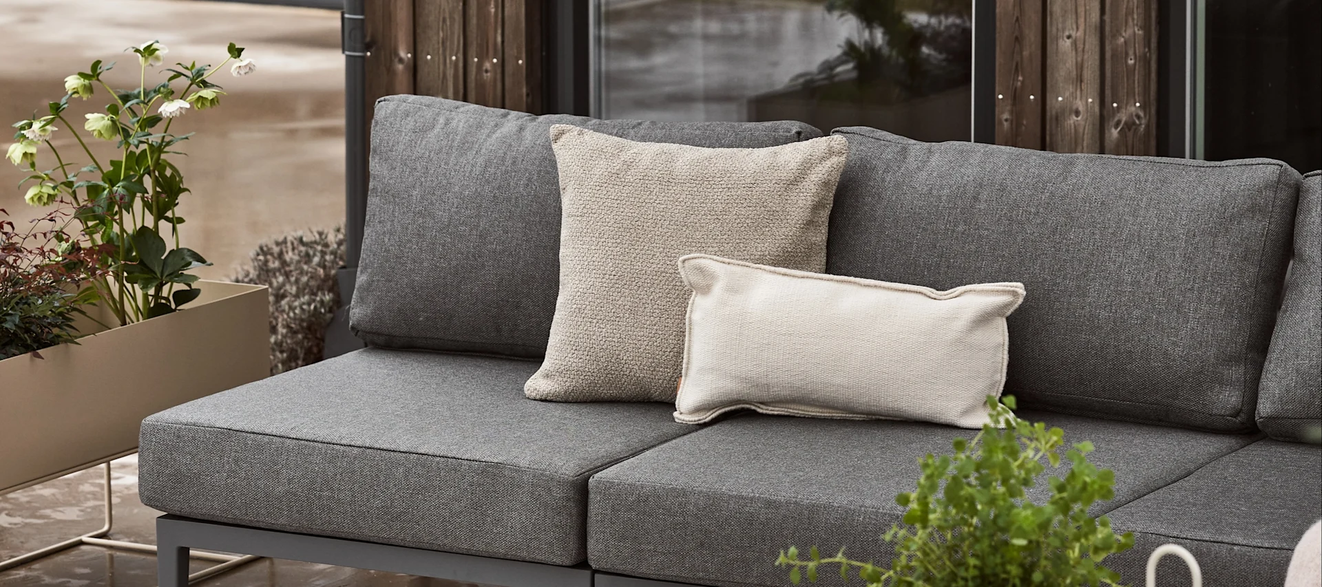 Ein graues Outdoor-Sofa mit zwei hellen Zierkissen, neben einem Pflanzgefäß mit grünen Pflanzen und weißen Blüten.