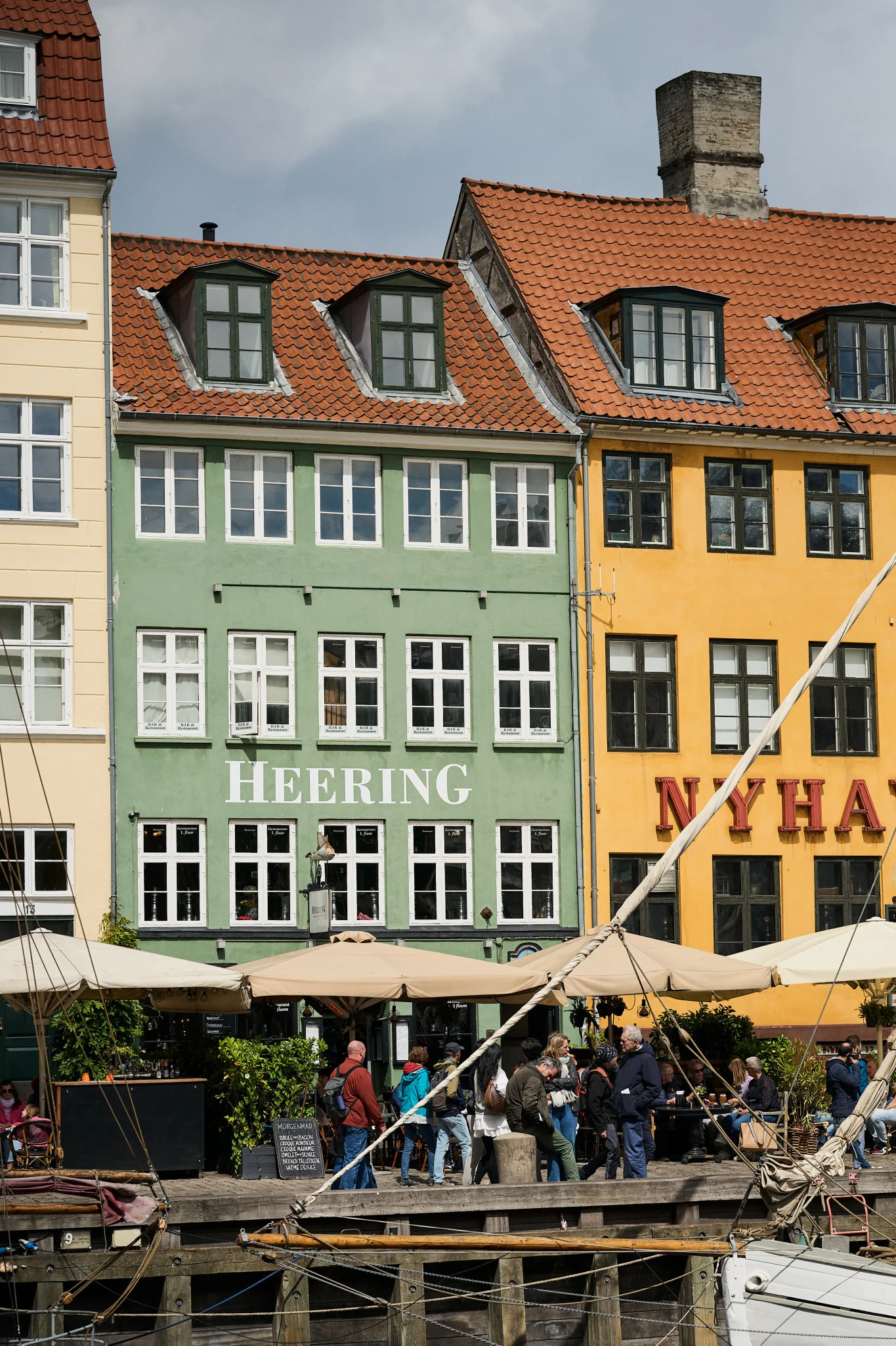 Lebhafte historische Gebäude mit roten Dächern säumen einen belebten Kanal. Menschen speisen in Straßencafés, mit Booten im Vordergrund.