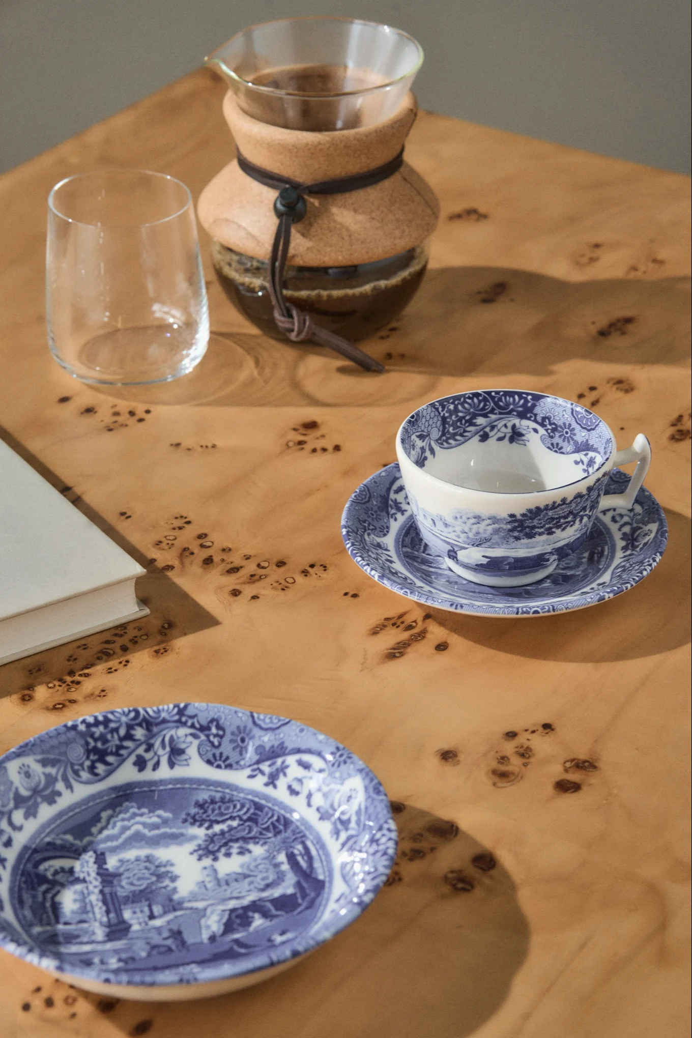 Eine Pour-over-Kaffeemaschine, ein leeres Glas, eine blau-weiß gemusterte Teetasse auf einer Untertasse und ein passender Teller auf einem hellen Holztisch.