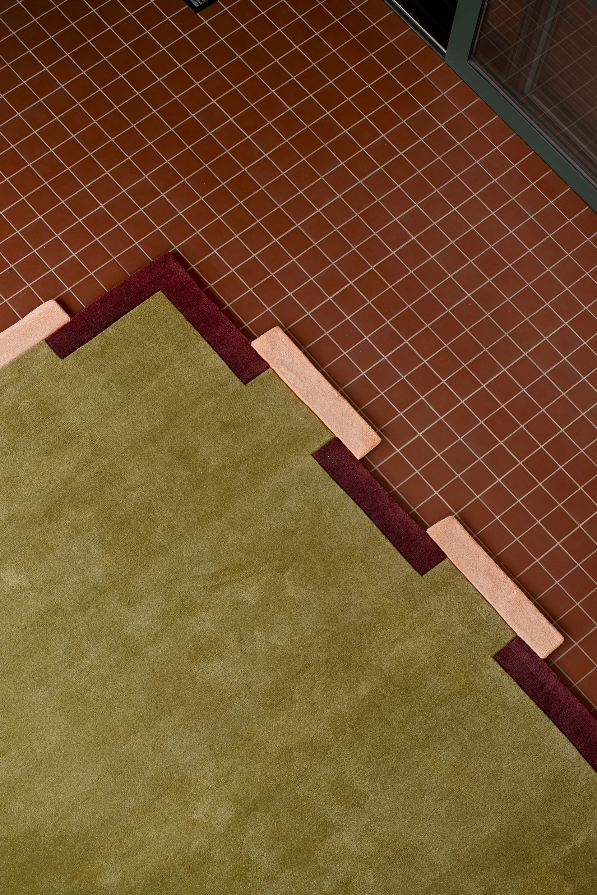 Ein geometrischer Teppich in Grün, Kastanienbraun und Pfirsich mit gestuftem Muster auf einem rotbraunen Fliesenboden.