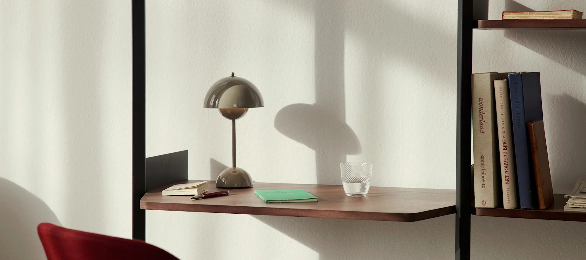 Eine moderne pilzförmige Schreibtischlampe, ein Glas Wasser und Notizbücher auf einem Holzschreibtischregal neben einem Bücherregal.