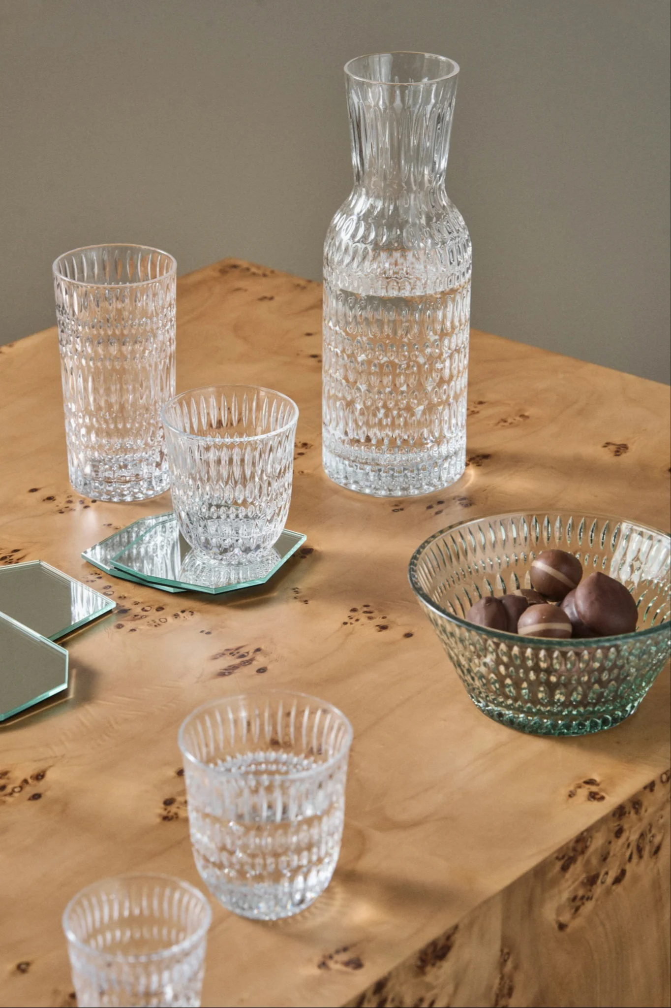 Karaffe, Gläser und Schale mit Pralinen aus strukturiertem Glas auf einem Wurzelholztisch mit Spiegeluntersetzern.