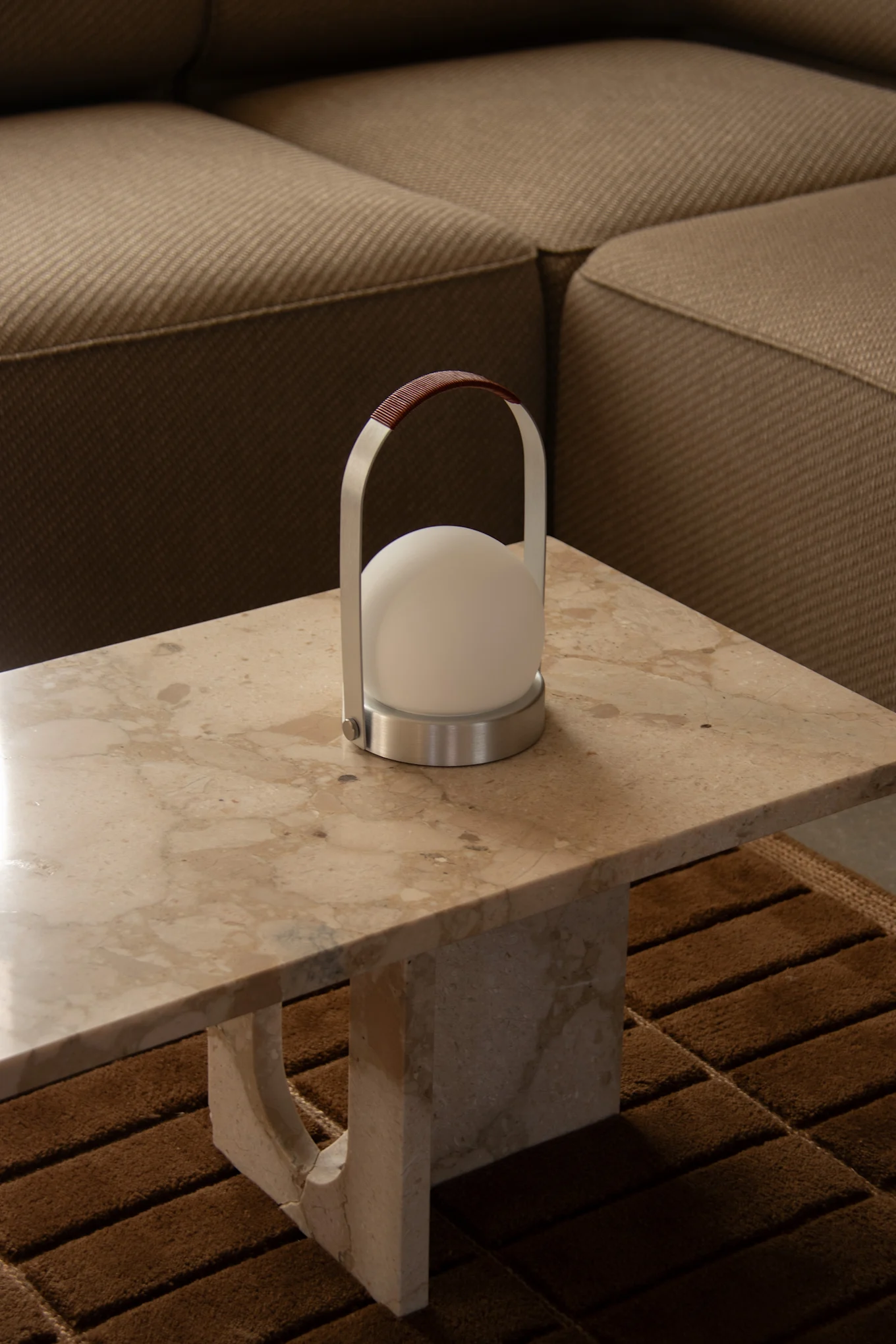 Eine moderne tragbare Lampe mit weißem Kugellicht, gebürstetem Metallrahmen und braunem Ledergriff steht auf einem hellen Marmortisch.