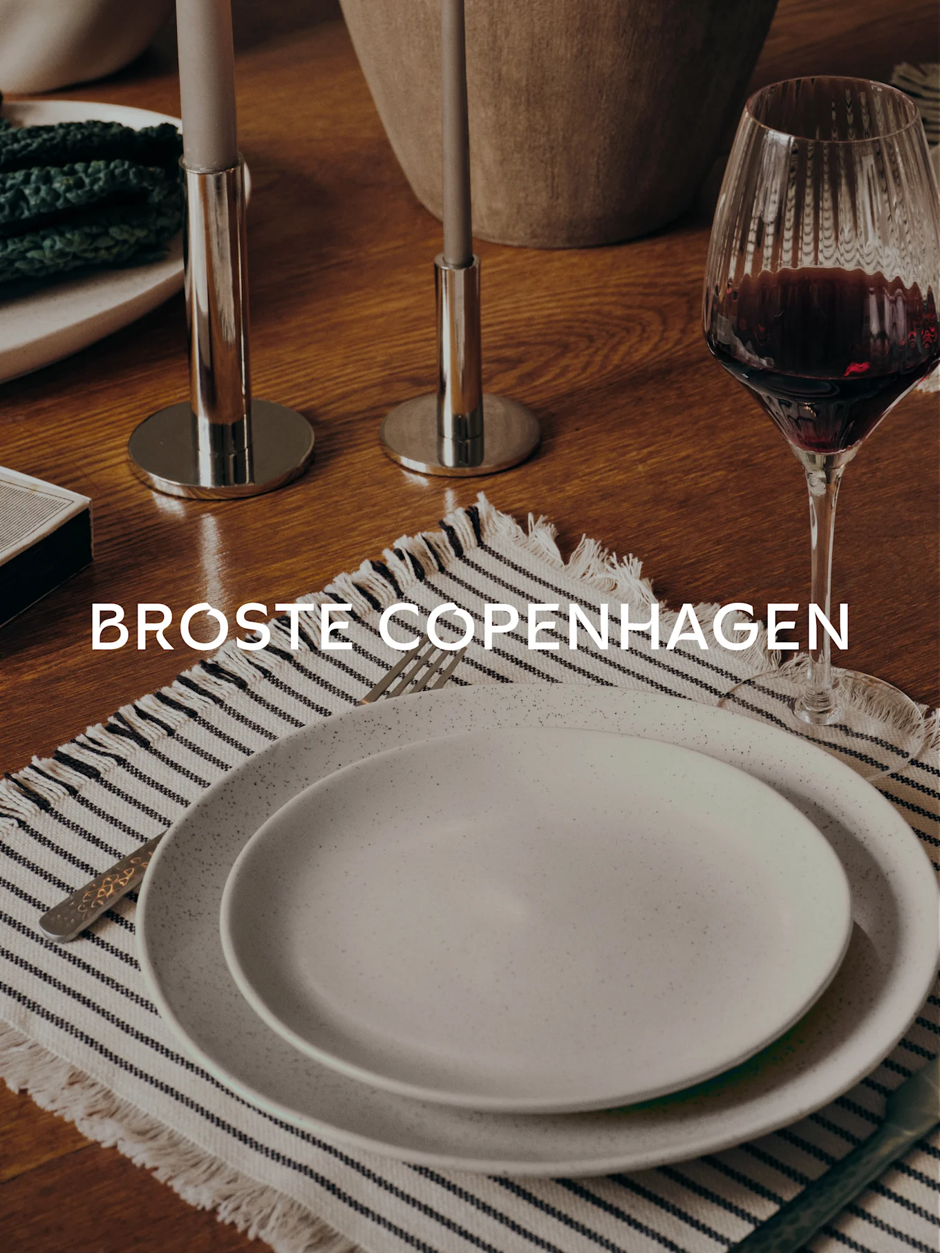 Esstisch mit zwei gesprenkelten Tellern auf gestreiftem Platzdeckchen, Gabel, Weinglas, zwei Kerzenhaltern und dem Text 'BROSTE COPENHAGEN'.