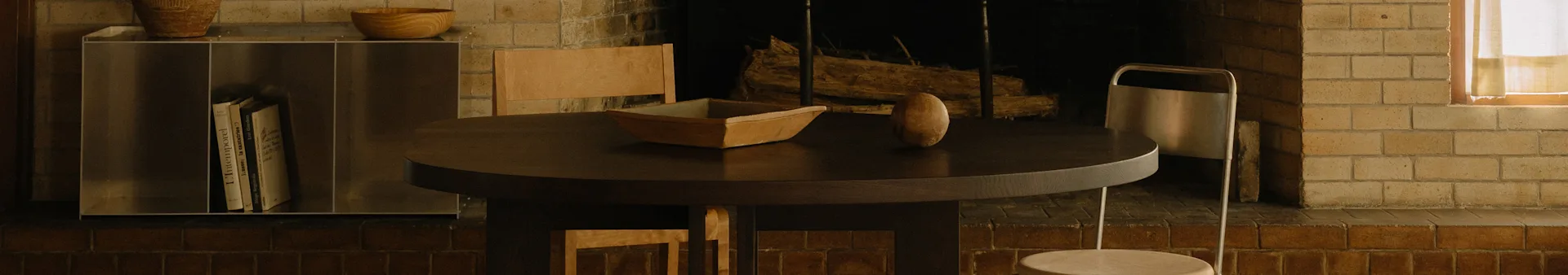 Ein gemütlicher Essbereich mit einem runden dunklen Holztisch, einer Holzschale, einem ovalen Objekt, Stühlen und einem Kamin.