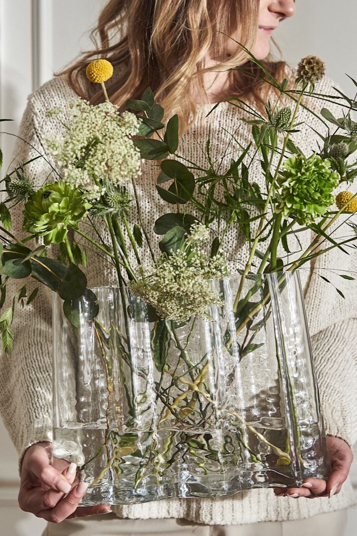 Eine in hellen Tönen gekleidete Frau hält die Gry Vase, welche mit wilden Frühlingsblumen gefüllt ist. 