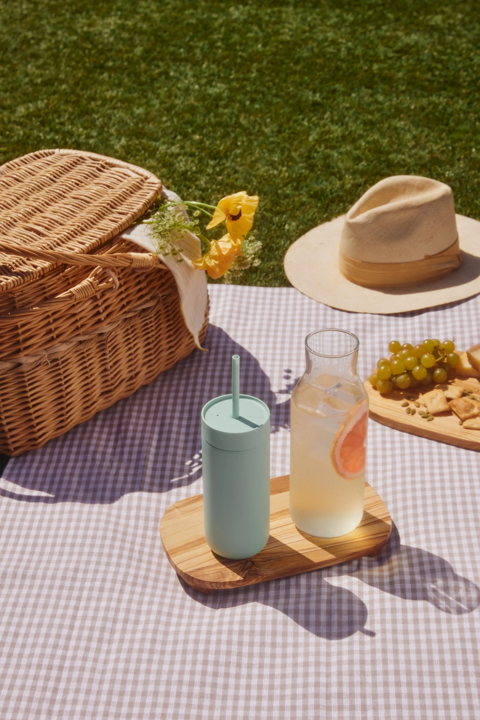 Picknick auf einer karierten Decke: Weidenkorb, hellgrüner Becher, Grapefruitgetränk, Trauben und Cracker auf Gras.